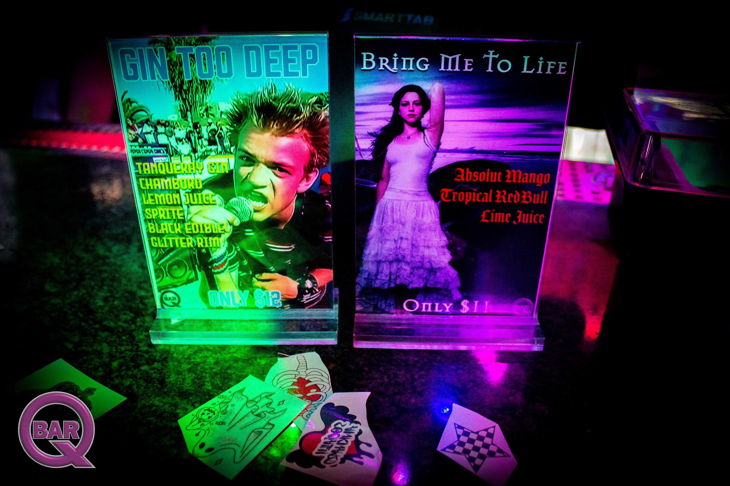 Neon-lit photo of two drink menus, one featuring a boy singing with the text 'GIN TOO DEEP' and another featuring a woman at the beach with the text 'BRING ME TO LIFE'. The scene includes various stickers and papers on a dark surface.