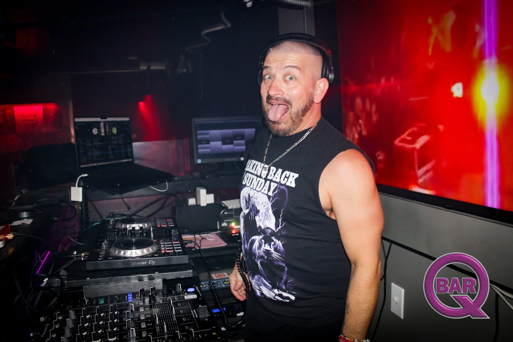A man with a beard and short hair wearing a black sleeveless shirt, headphones, and a chain necklace, standing behind a DJ booth with DJ equipment, in a nightclub with red lighting and a screen in the background.