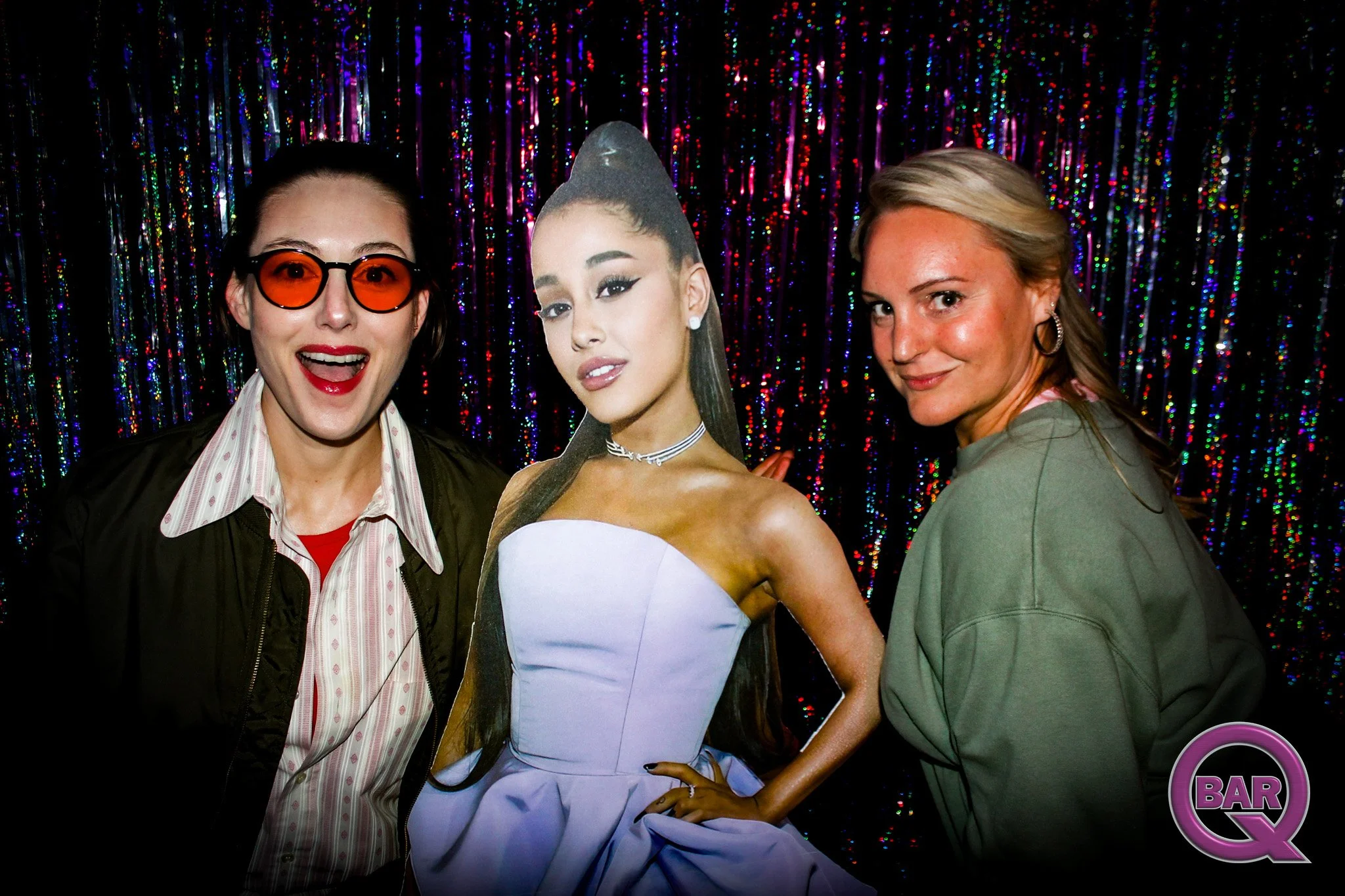 Two women stand beside a life-sized cardboard cutout of a woman in a white dress with a high slit, set against a shiny, colorful, striped backdrop. One woman wears glasses with orange lenses, a striped shirt, and a dark jacket. The other woman has bl