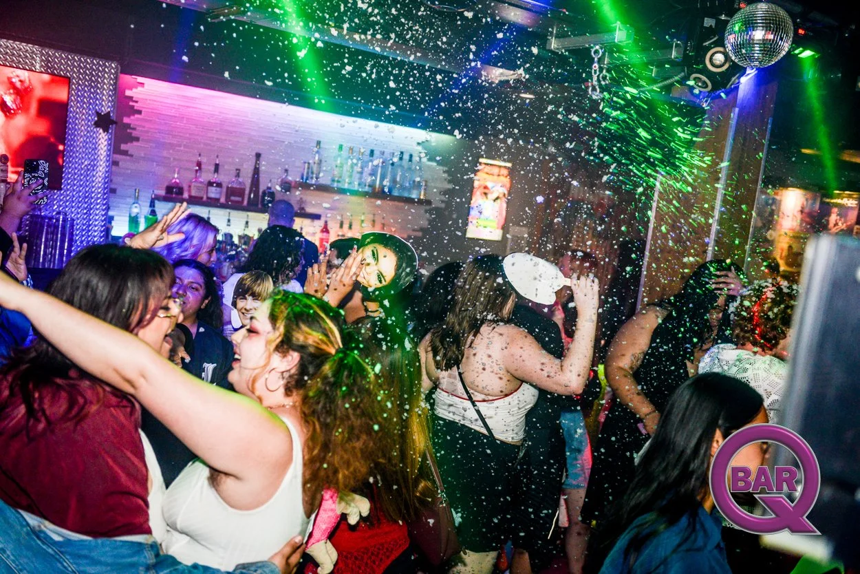People dancing and enjoying themselves in a nightclub with colorful lights, a disco ball, and foam falling from the ceiling.
