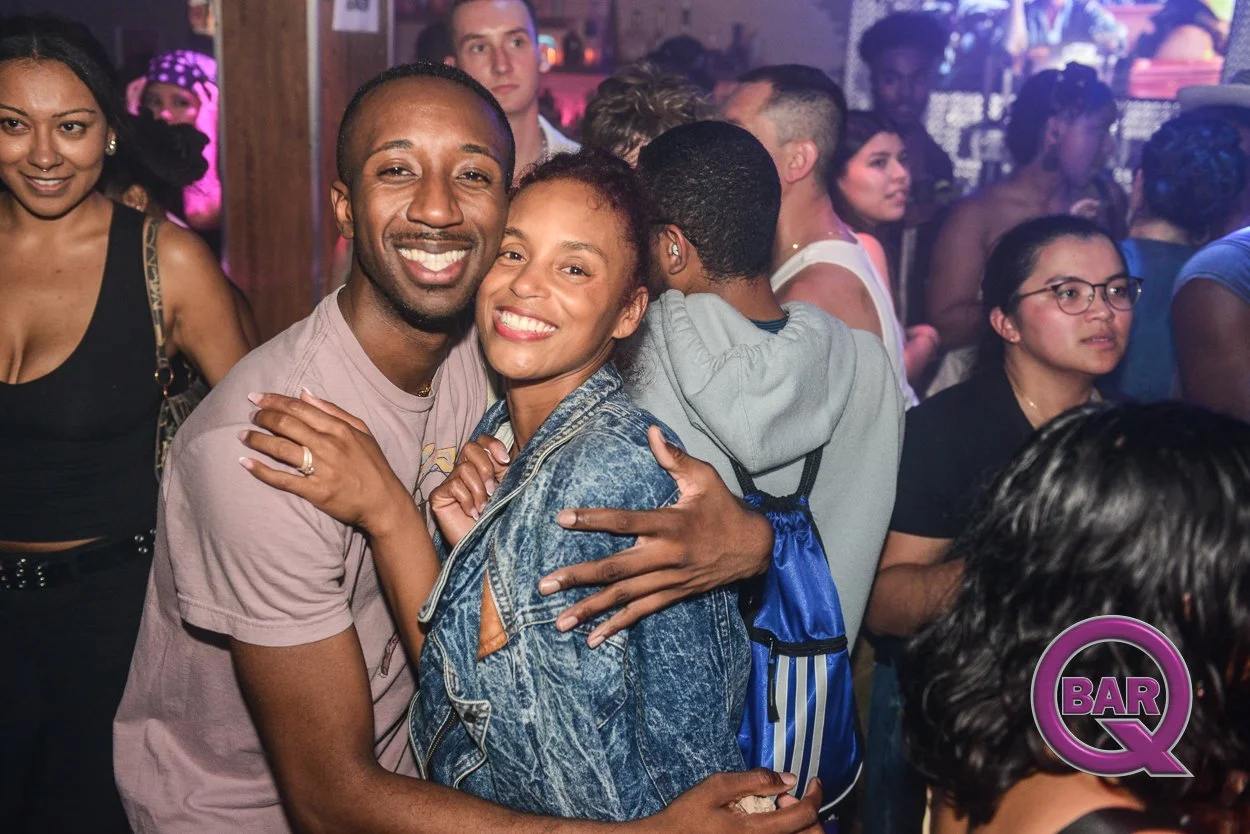 Group of smiling people hugging and dancing at a nightclub, with colorful lights and a bar in the background.