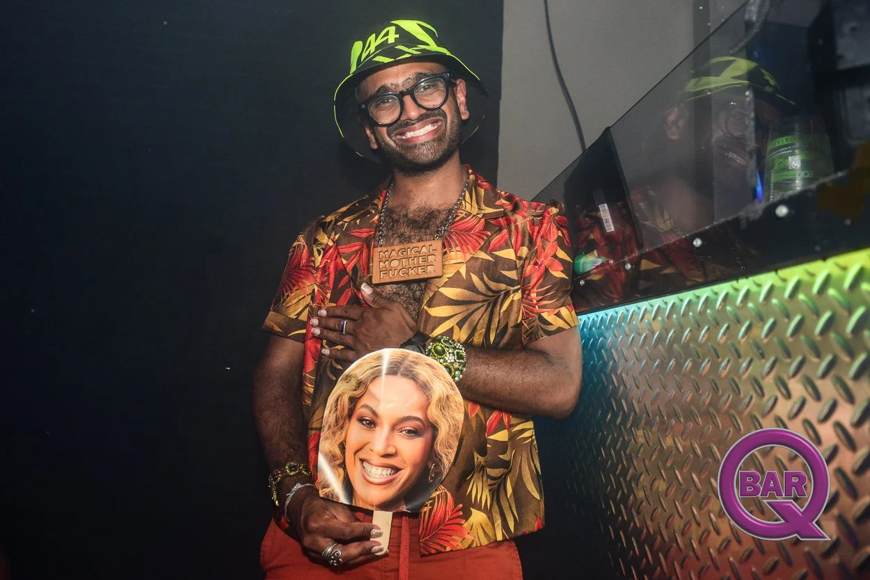 Man in a colorful floral shirt, wearing a hat and glasses, smiling at a party or club. He holds a photo of a woman smiling. There is a large wooden necklace with the words "Magical Mother Fucker." He has various jewelry and a wristwatch. The backgrou
