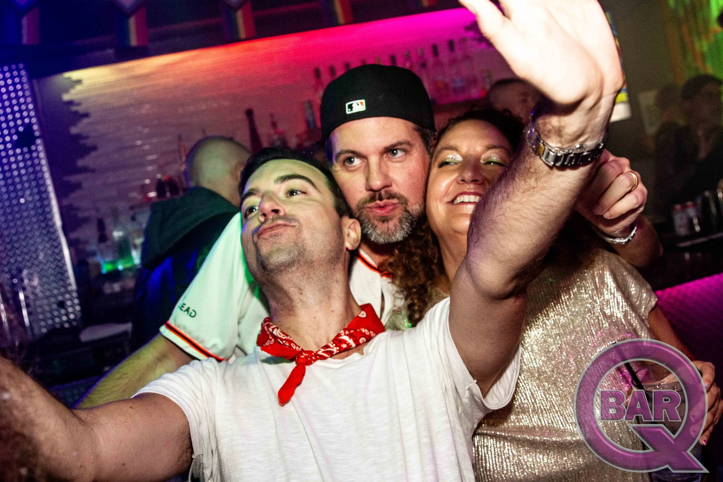 Three friends taking a selfie at a bar or nightclub, with colorful lighting in the background. The person on the left wears a white shirt with a red bandana, the middle person has a beard and wears a black cap, and the person on the right is smiling 