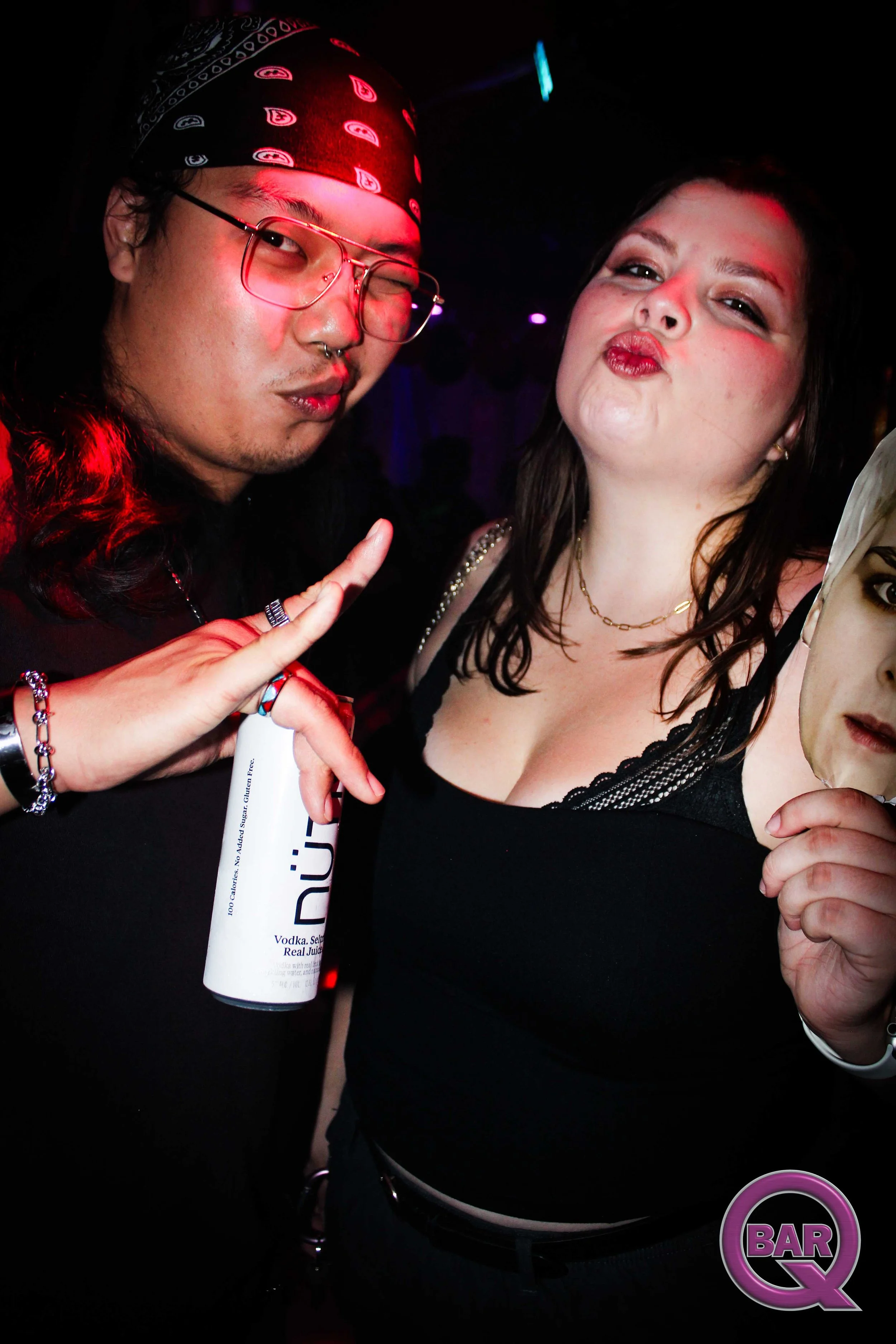 Two people posing in a dark setting with colorful lights. The person on the left, wearing a bandana, glasses, and jewelry, is holding a vodka bottle. The person on the right, with dark hair and makeup, is making a kiss face. In the bottom right corne