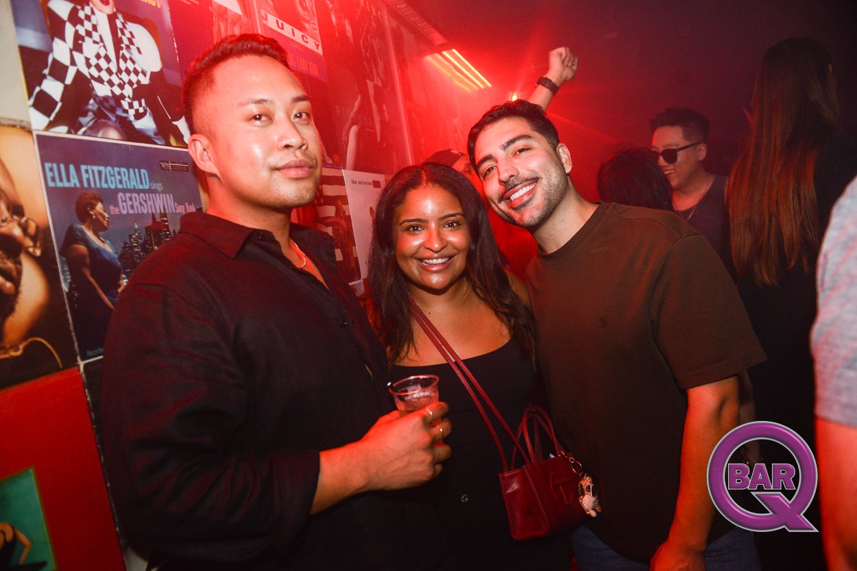 Three friends smiling and partying in a nightclub with red lighting, posters on the wall, and people in the background. One man is holding a drink, and the women are dressed casually.