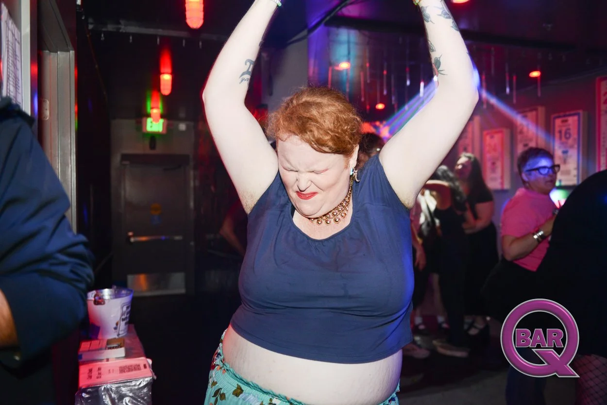 A woman with red hair dancing with her eyes closed at a nightclub, wearing a blue top and a necklace, with other people dancing in the background and colorful lights illuminating the scene.