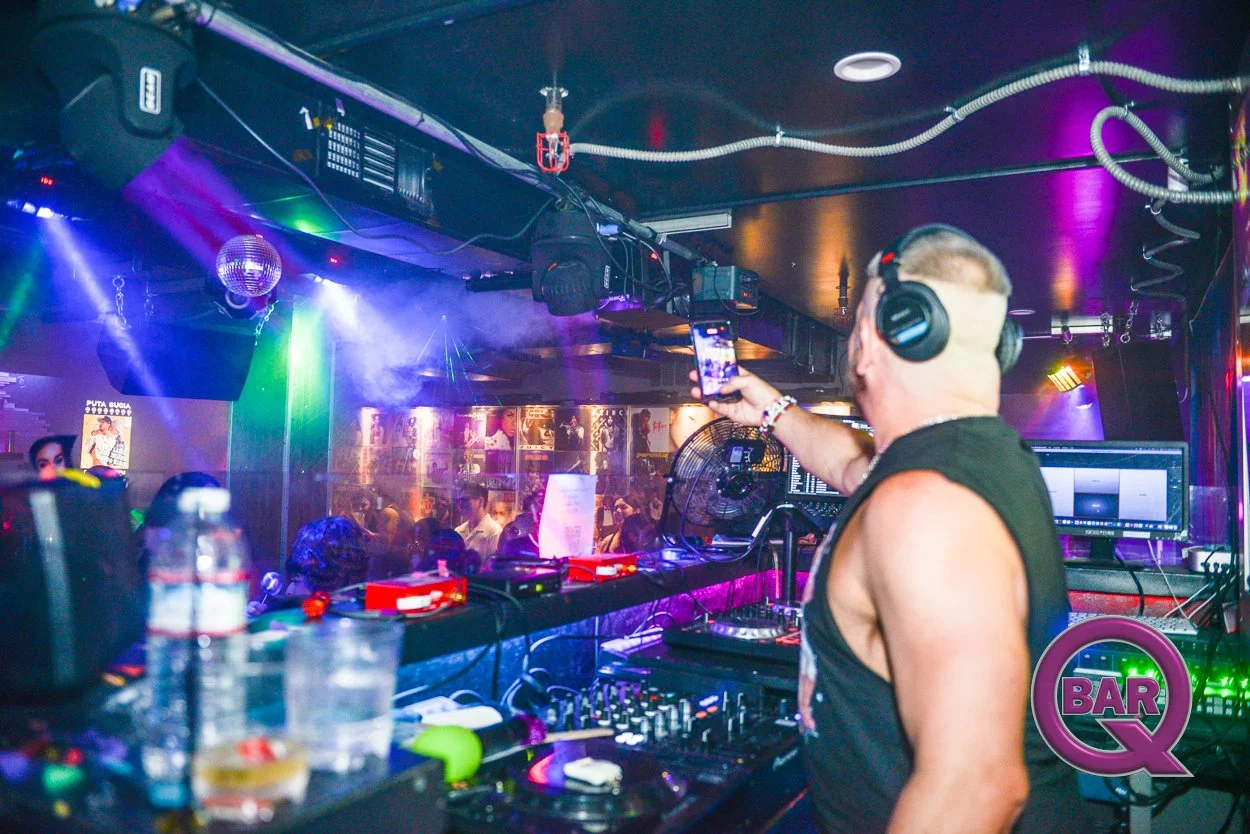 A DJ wearing headphones and a sleeveless shirt is mixing music at a nightclub. The club has colorful lighting, a disco ball, and a crowd of people dancing in the background.