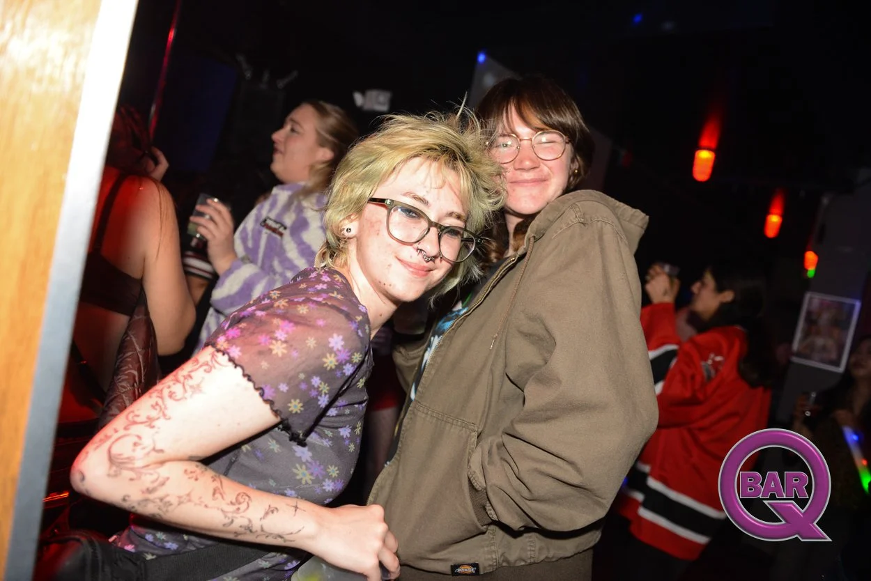 Two young women smiling together at a bar, one with glasses and tattoos, and the other with glasses and a brown jacket, with other people in the background.