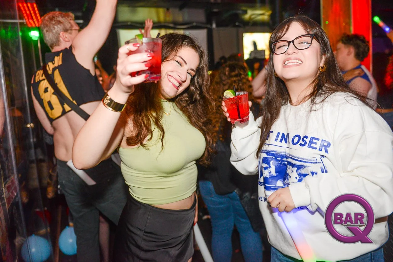 Two young women celebrating with red cocktails in a lively bar or nightclub with colorful lights and other people in the background.
