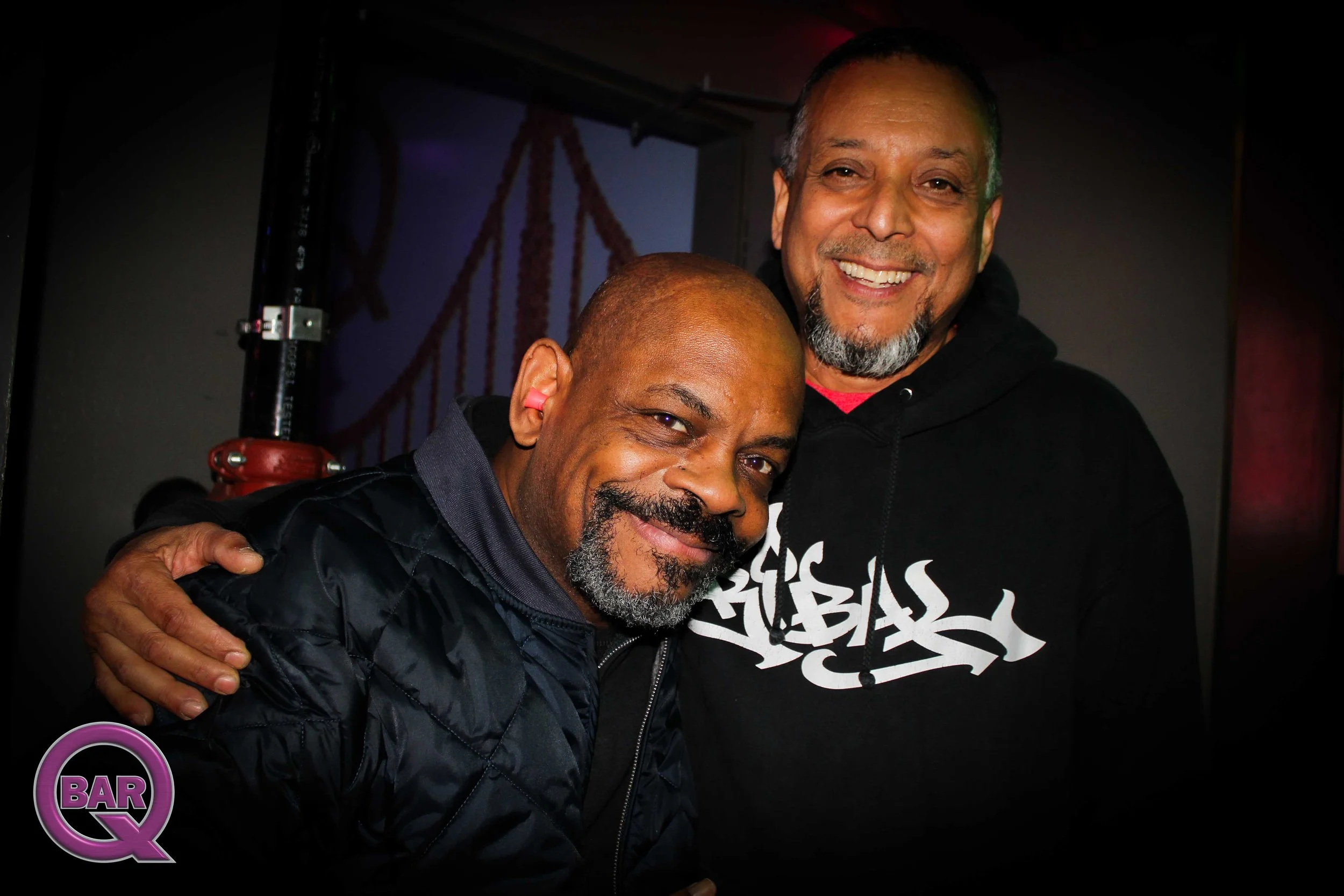 Two smiling men with facial hair embracing at a bar or nightclub, with a dark background and purple and red lighting effects.