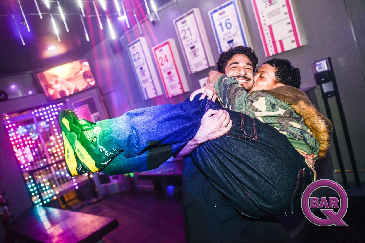 Two men are happily embracing in a bar or nightclub with colorful neon lights and formatted graphics on the wall.