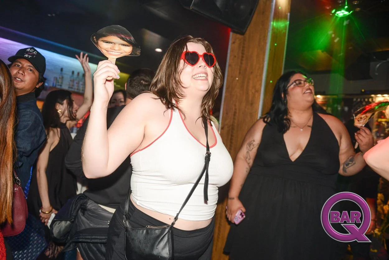 People dancing and having fun at a bar. A woman in the foreground is wearing red heart-shaped sunglasses and holding a photo mask of a woman's face. The background shows other people enjoying the lively atmosphere with colorful lights and a bar area.