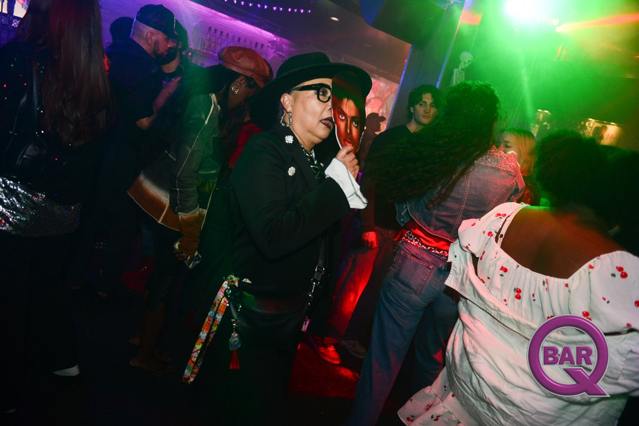 People dancing and socializing in a nightclub with colorful lighting, including a woman with dark makeup, glasses, and a black hat, holding a tissue near her face.