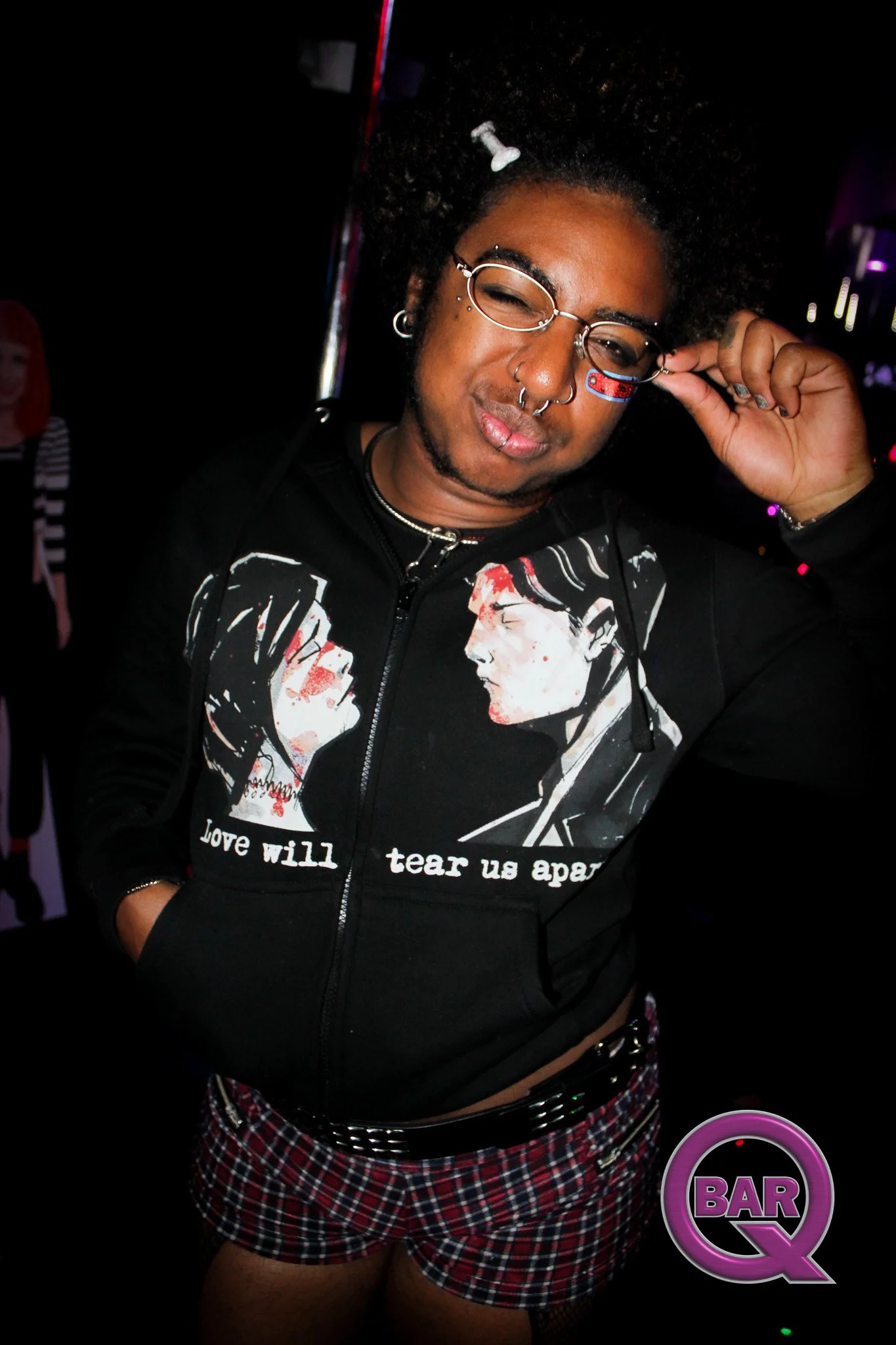 A person with curly hair and glasses adjusting their sunglasses inside a club or bar with dark lighting and neon signs.
