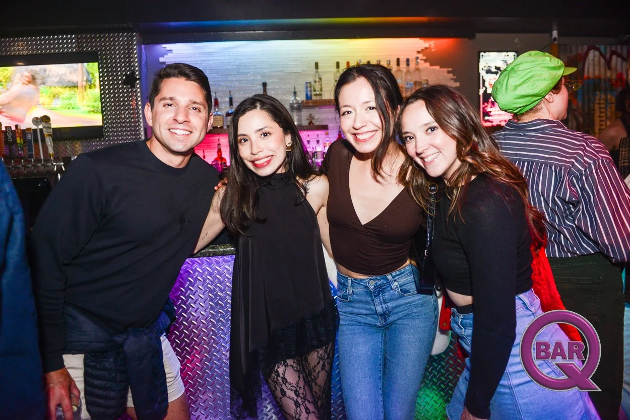 Four young adults smiling and posing together in a bar with colorful lighting and a bar counter behind them.