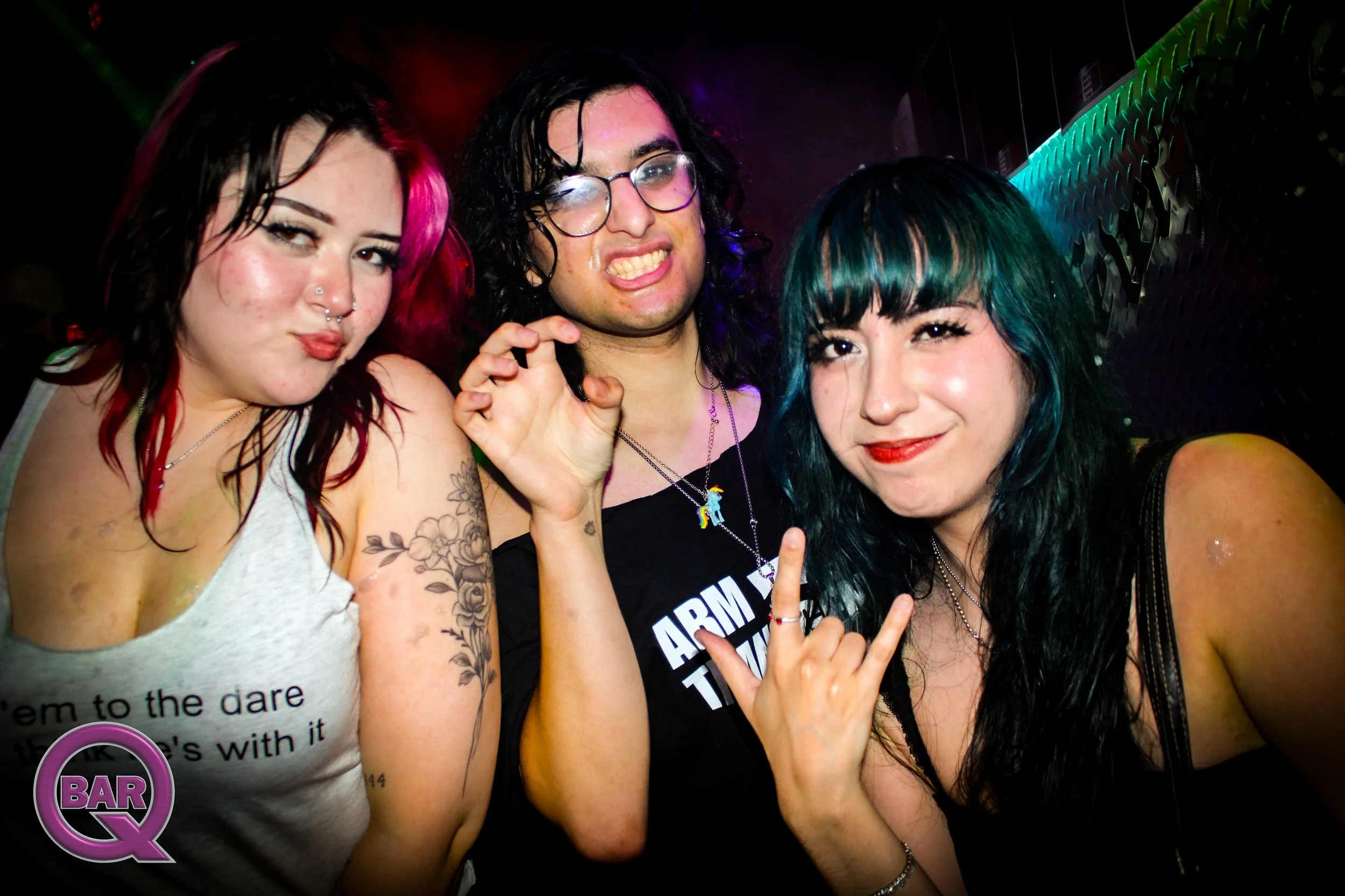 Three women at a nightclub or party, smiling and making gestures for the camera. The woman on the left has tattoos, piercings, and red and black hair. The woman in the center wears glasses and has dark, wavy hair. The woman on the right has dark hair