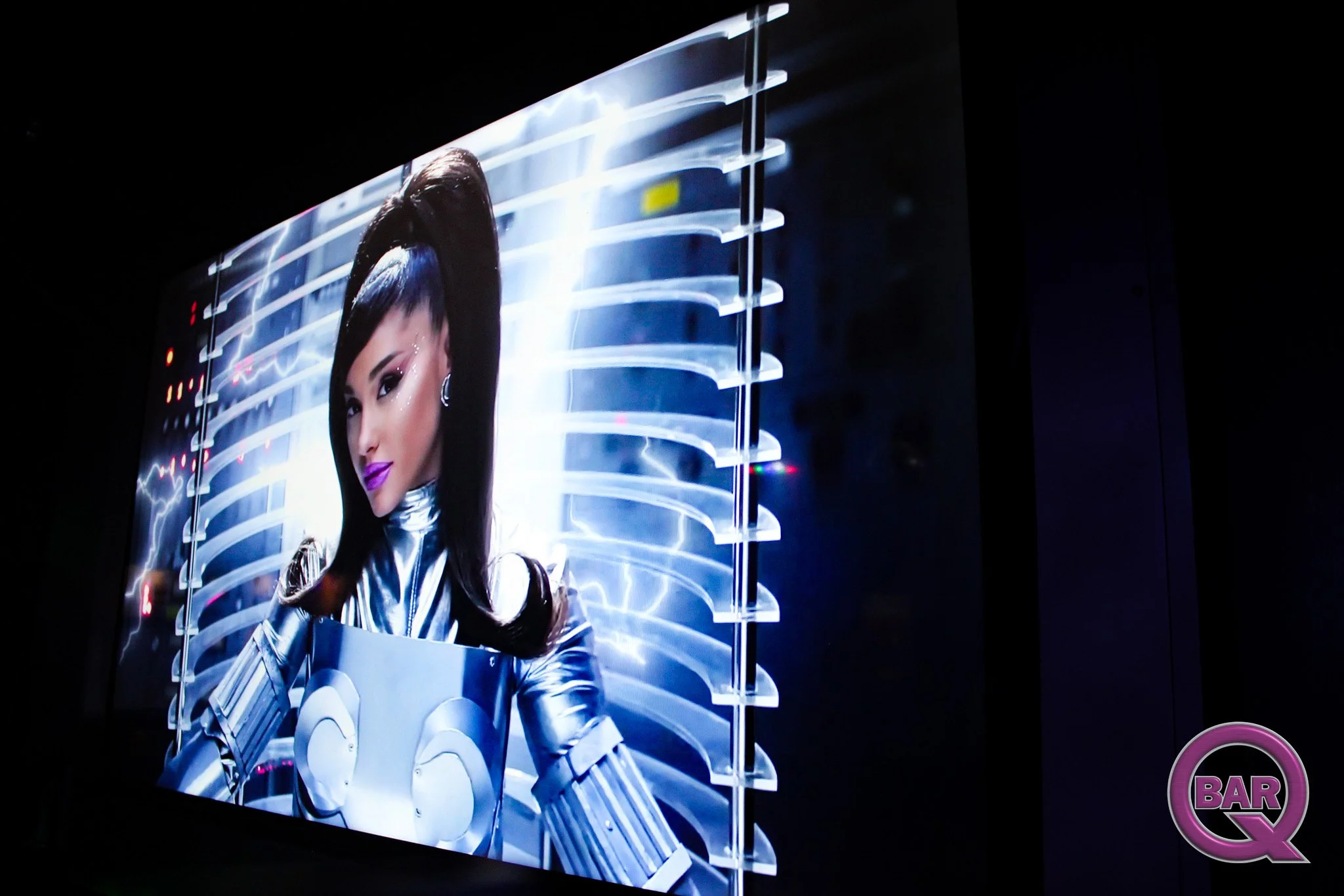 A woman with long dark hair and purple lipstick dressed in a futuristic silver and blue suit, standing in front of a science fiction-inspired backdrop with curved glowing lines and a lightning effect.
