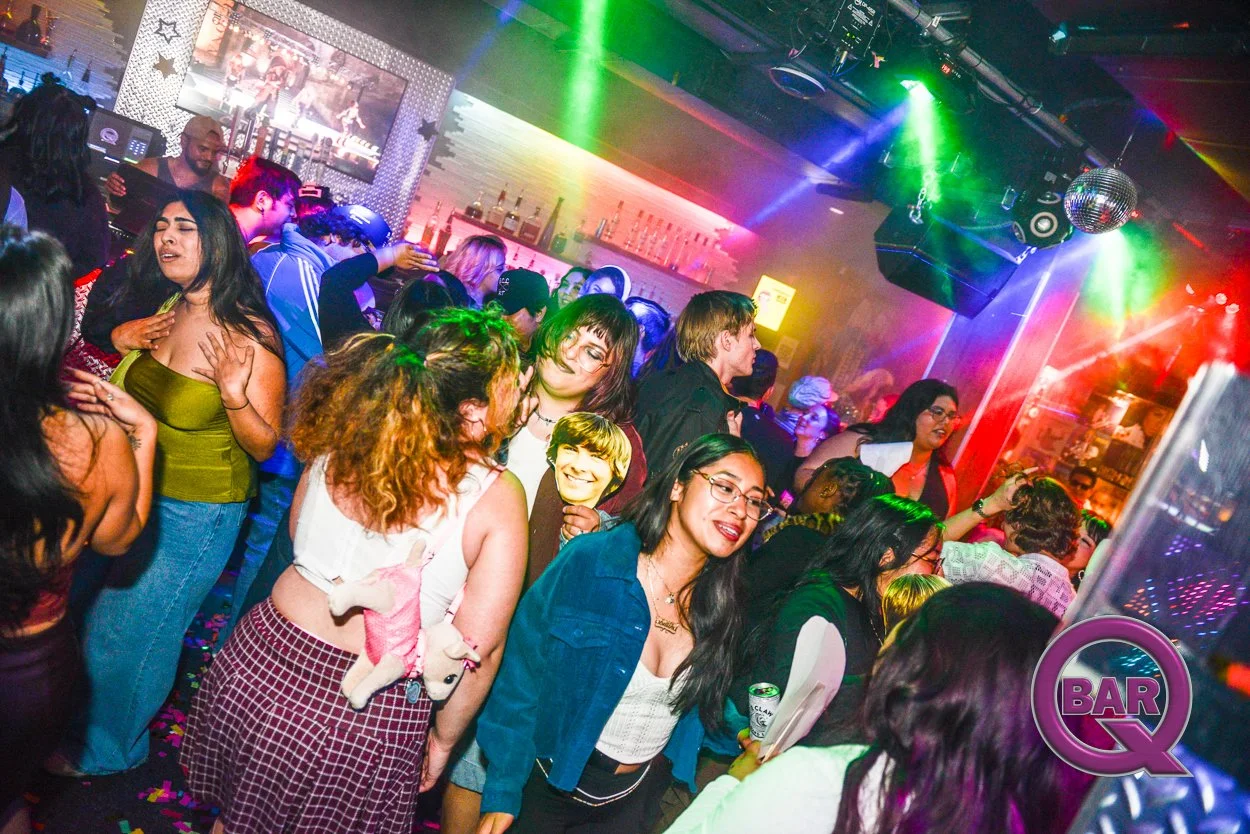 People dancing and socializing in a colorful nightclub with rainbow lighting and a DJ booth in the background.