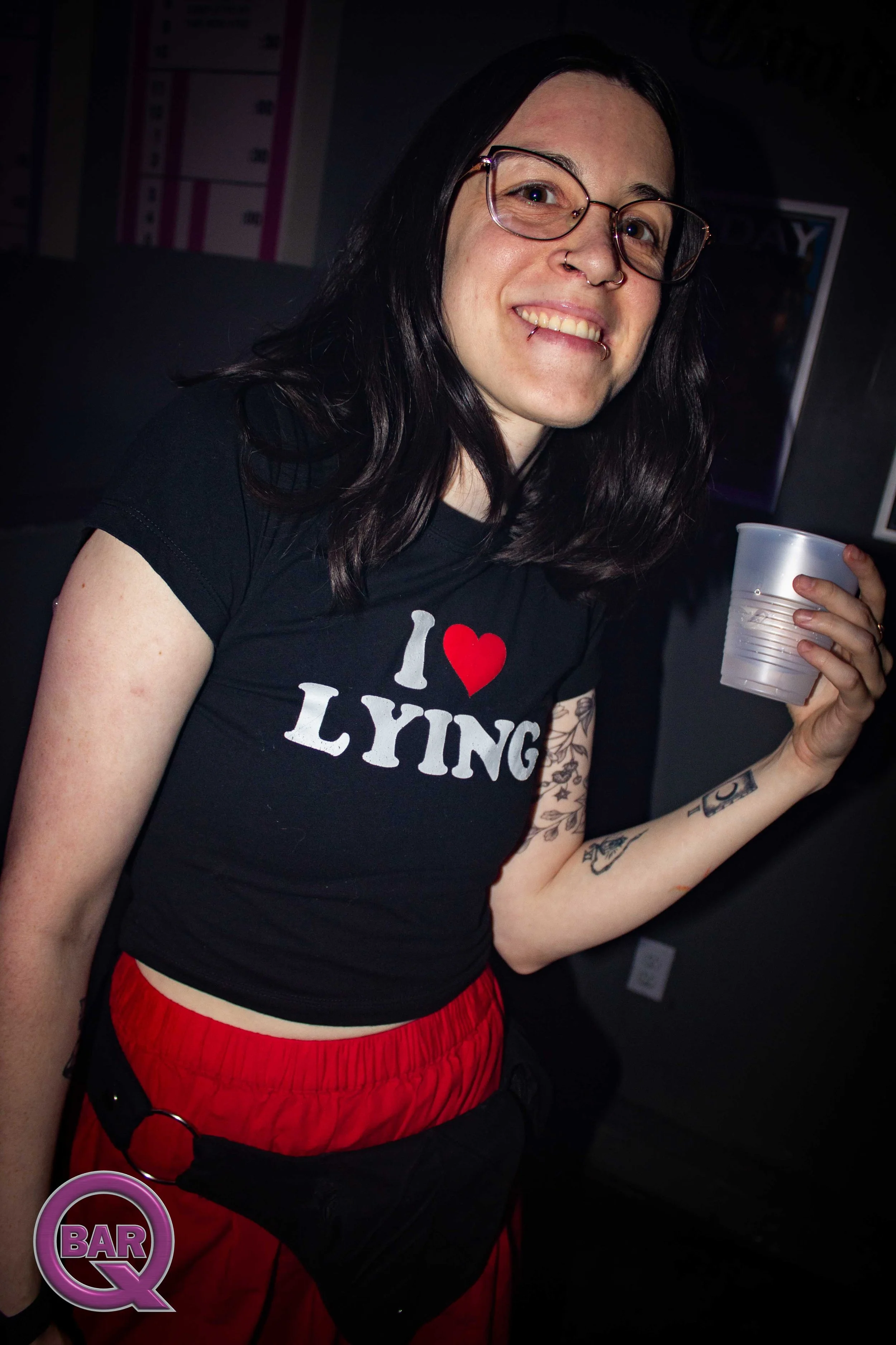 A woman smiling, holding a clear plastic cup, wearing a black t-shirt with the phrase 'I ♥ LYING' and red shorts, standing in a dimly lit room with posters on the wall.