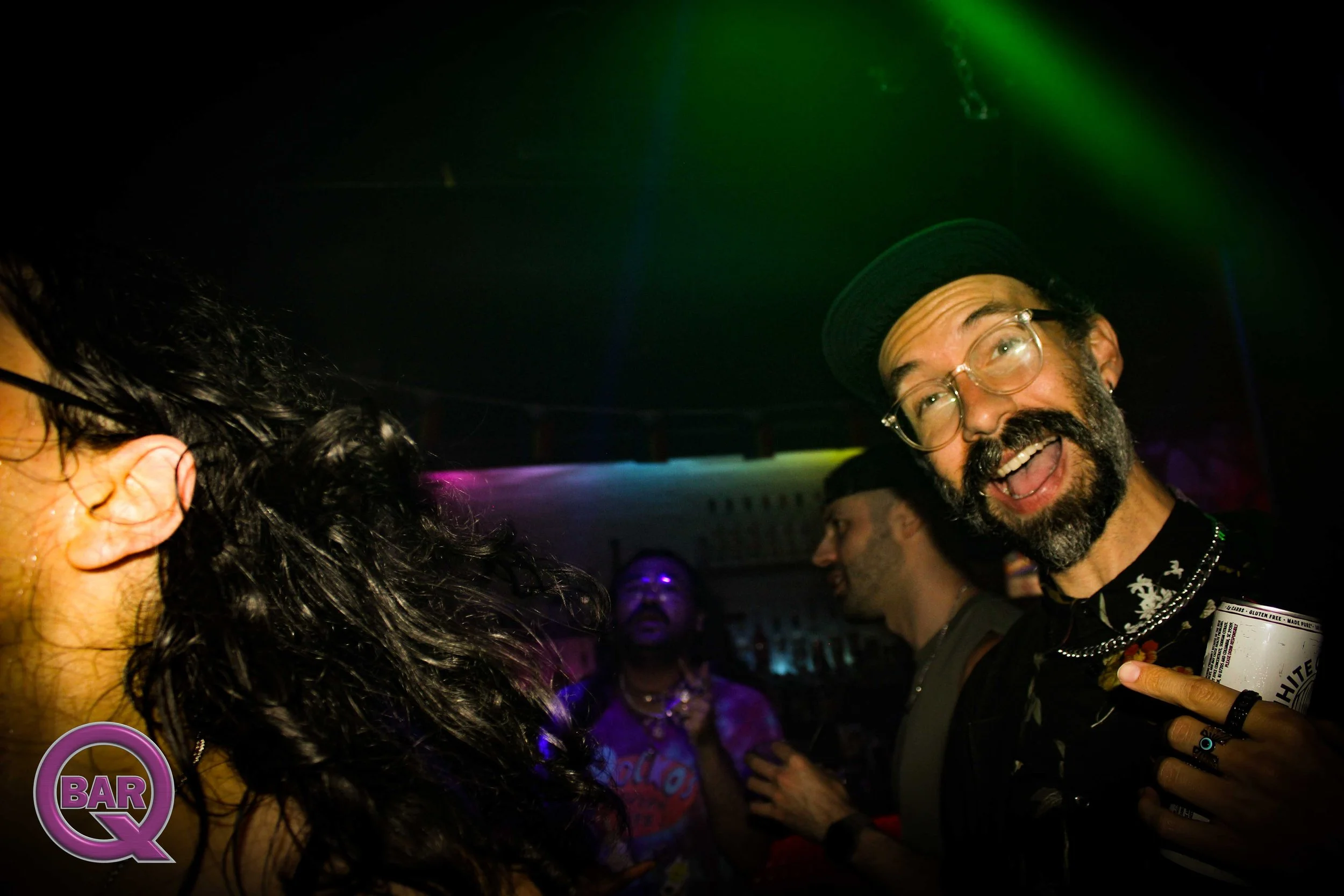 A group of people socializing at a bar in a dimly lit setting with colorful lights. One man with a beard, glasses, and a hat is holding a bottle and smiling.