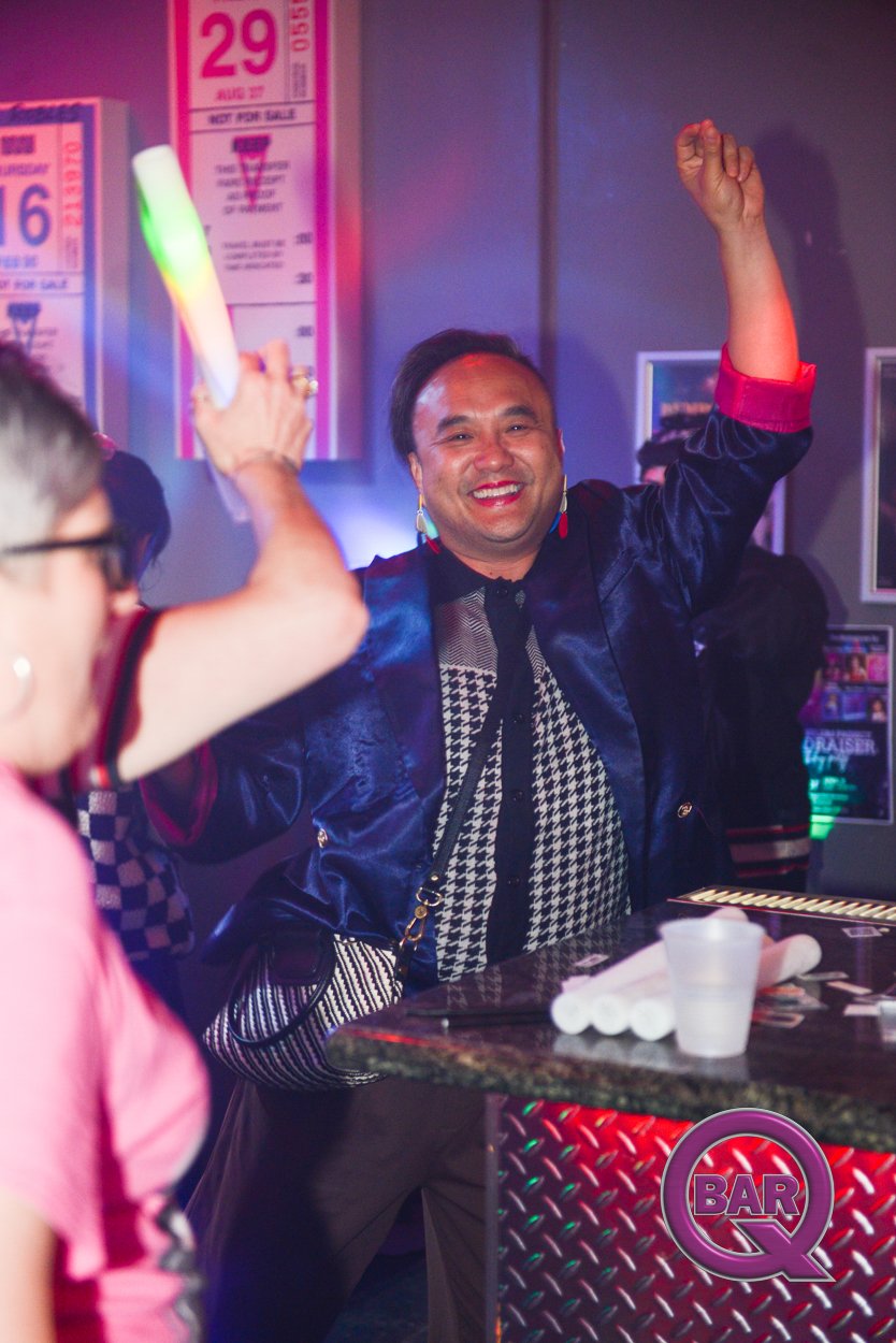 Happy man with short dark hair smiling and raising his right fist in a lively bar or nightclub, with colorful lighting and blurred background of other people and posters.