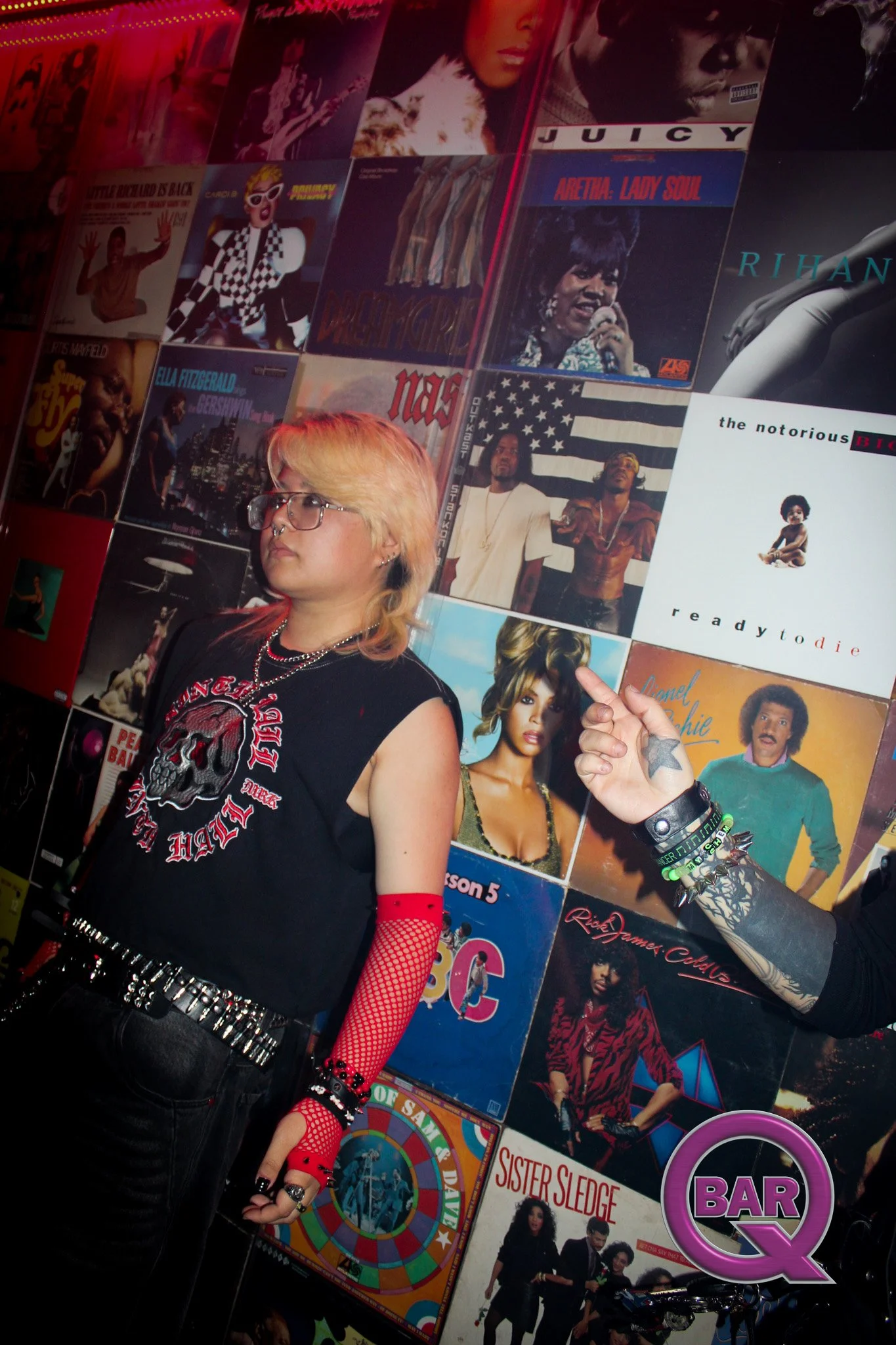 A young person with blonde hair, glasses, and punk style clothing standing in front of a wall covered with colorful album covers, including Aretha Franklin, Sister Sledge, and others. The person is wearing a black T-shirt with a skull design, red fis