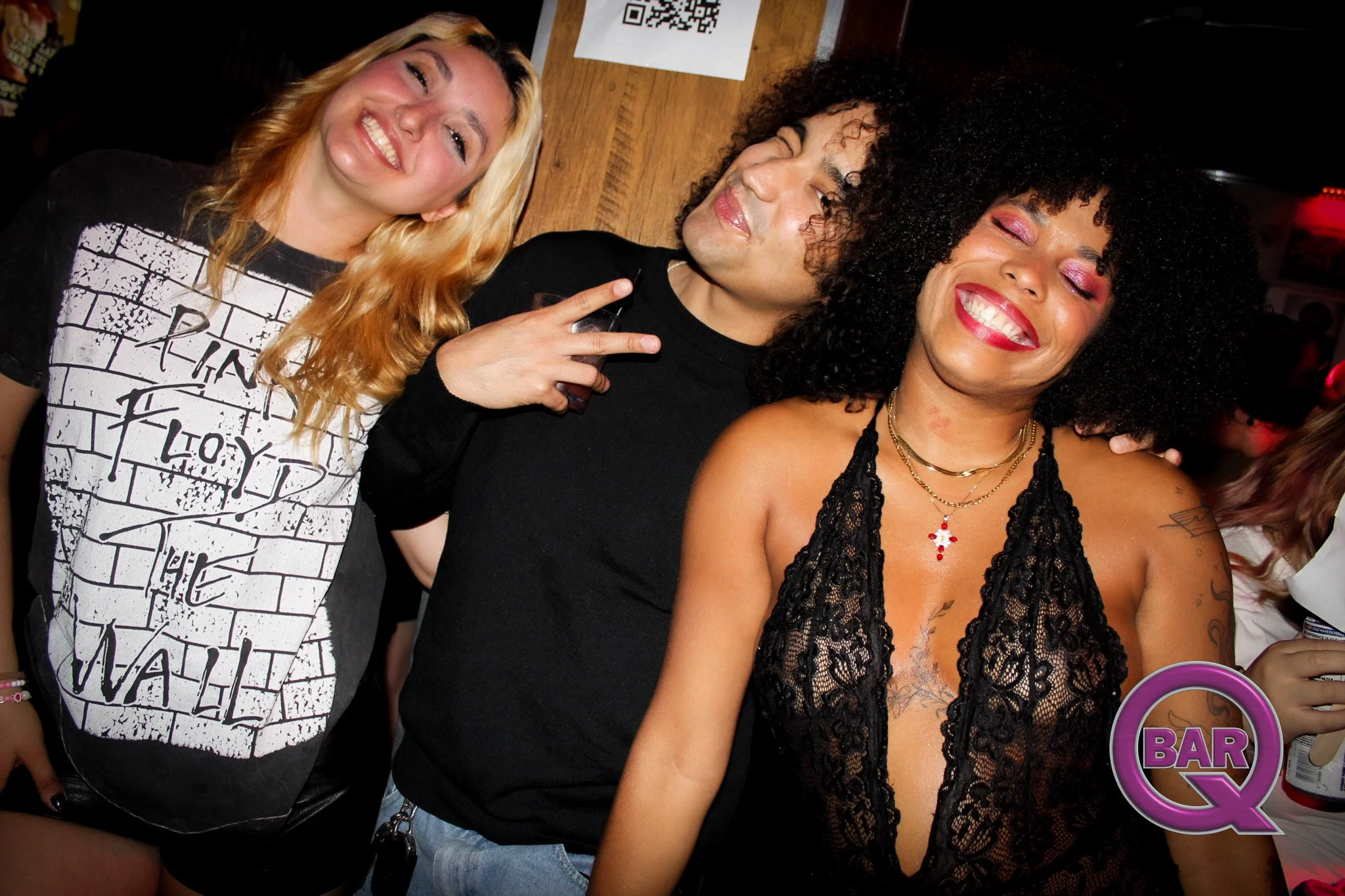 Three women smiling and posing closely together at a bar or club, with one woman wearing a black lace top, the middle woman with curly hair holding a drink, and the woman on the left with blonde hair wearing a graphic T-shirt.