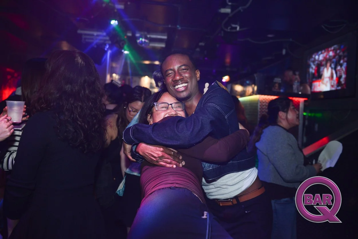 Two people hugging and smiling in a crowded nightclub with colorful lights and a television screen in the background.