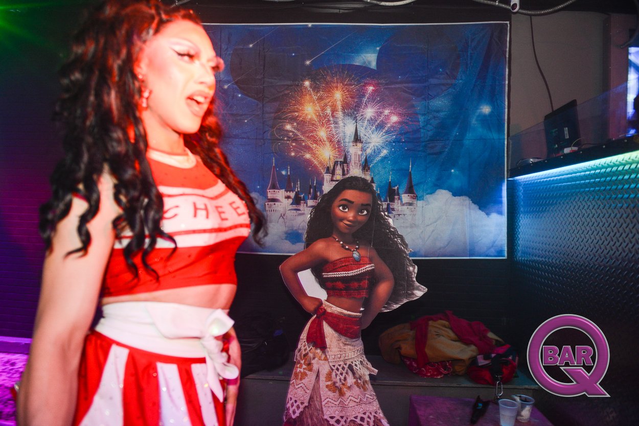 A woman singing or performing on stage, with a backdrop of a castle and fireworks, and a cardboard cutout of an animated woman in a tropical outfit in front of her. The scene is inside a bar or club, with a logo that says 'BAR Q' in the bottom right 