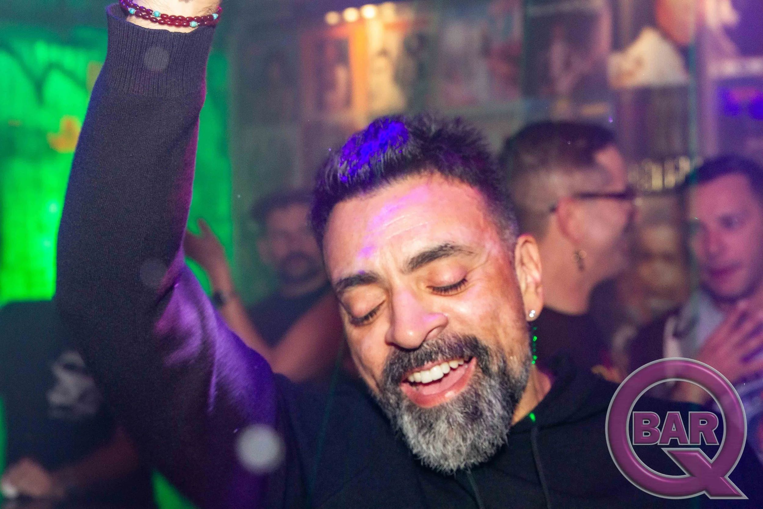 A man with a beard and mustache, smiling widely with closed eyes, at a social gathering or nightclub. He has dark, short hair and is wearing a dark shirt. There is a woman next to him with her arm raised, wearing a long-sleeve top. The background is 