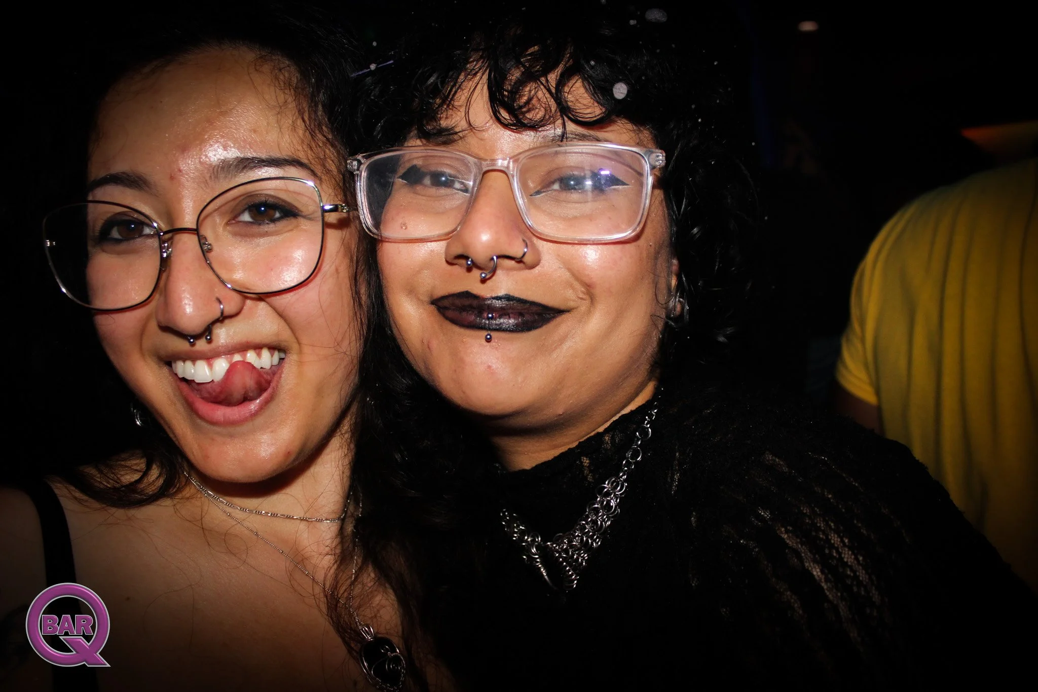 Two women with dark hair and glasses smiling at a night event, one with black lipstick and a septum piercing, the other with glasses and a wide smile, standing closely together.