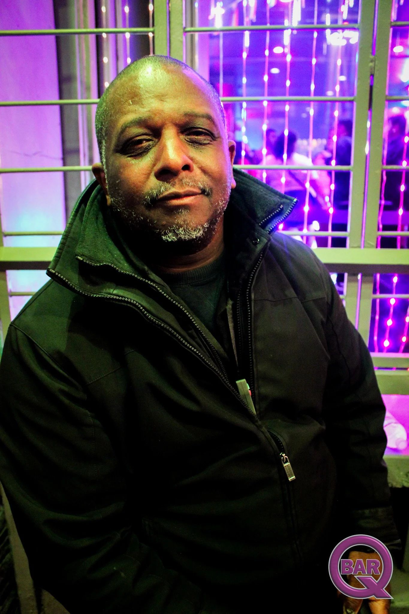 A middle-aged man with a shaved head, many grey facial hairs, and dark skin, smiling slightly. He is wearing a black jacket with a zippered front. The background features purple and blue neon lights and a metal grid, with people visible in the distan