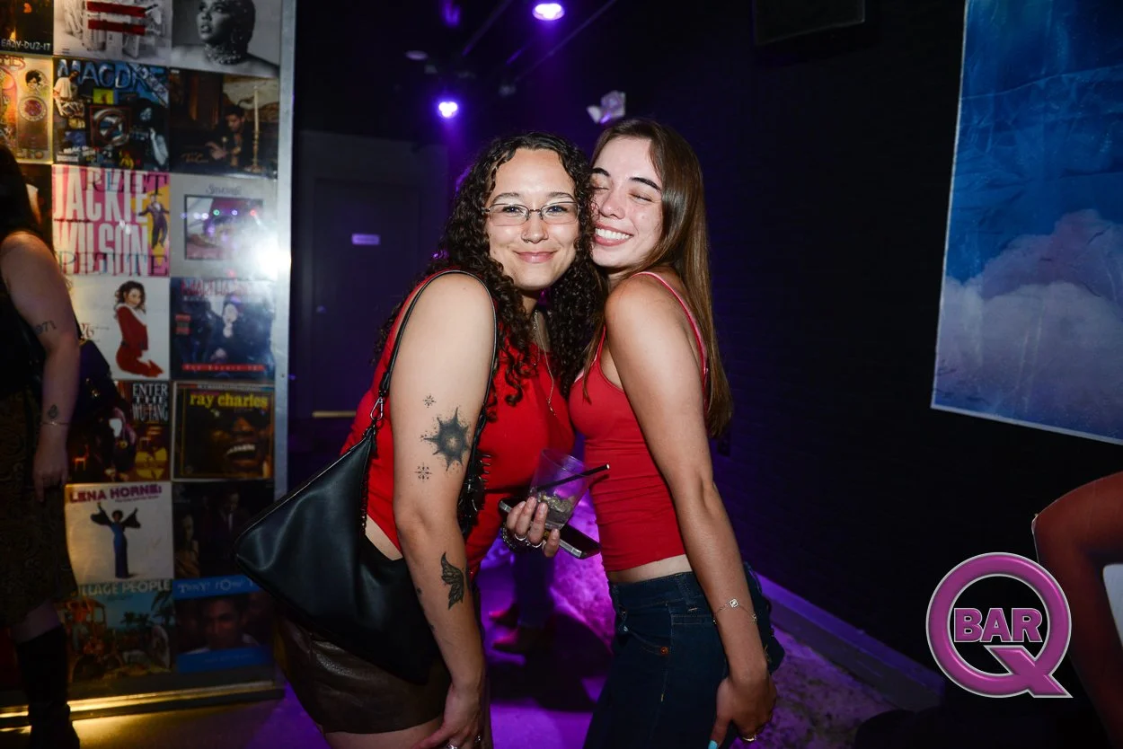 Two women in red tops and jeans hugging and smiling in a bar with posters on the wall and purple lighting.