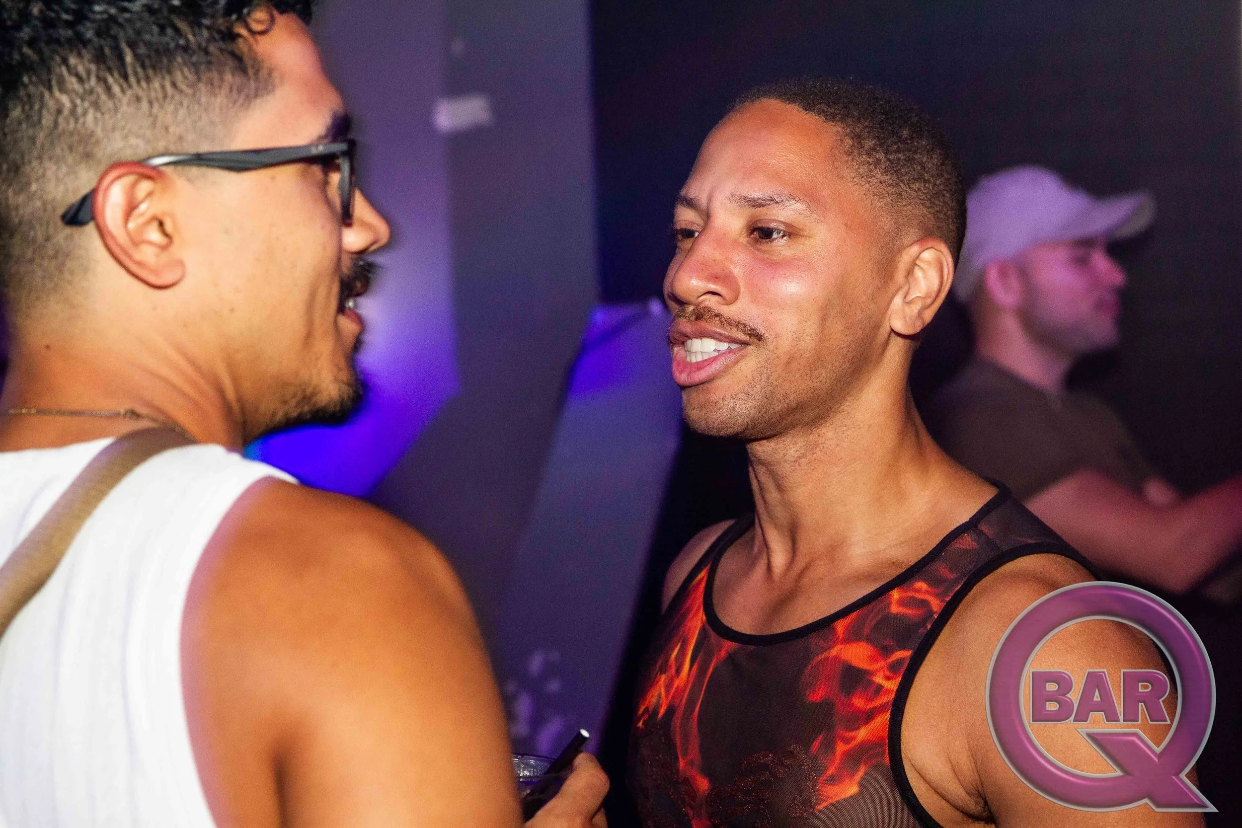 Two men in a bar or nightclub, facing each other closely, one smiling and the other looking serious. Background with purple lighting and other people.