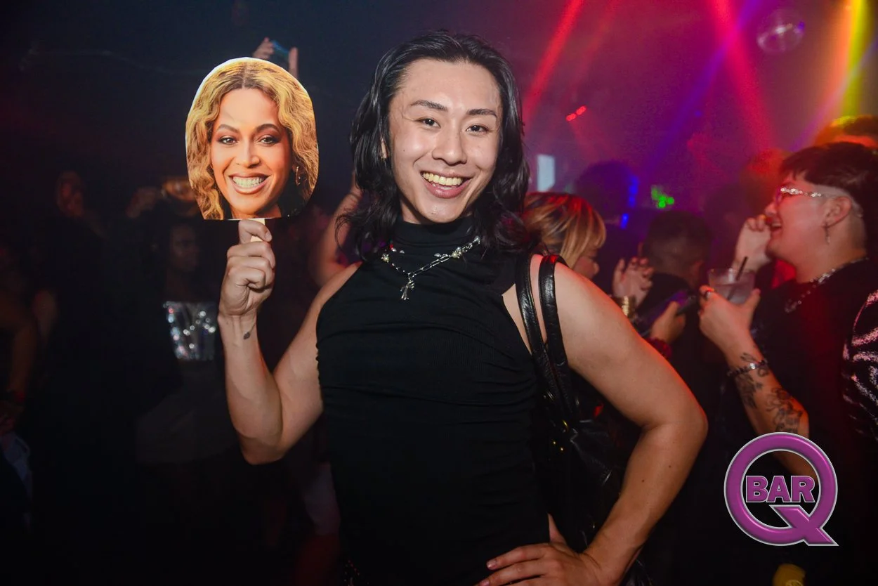 A person with long black hair smiling and holding a cutout picture of a woman with blonde curly hair at a crowded gay bar or nightclub with colorful lights and other people in the background.