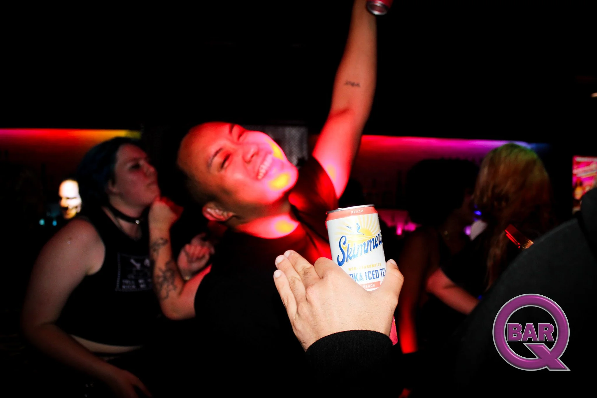 A woman with short hair smiling and raising her arm in a dark bar or club, illuminated by colorful lights, while someone holds a can of Skimmed peach-flavored sparkling water toward her.