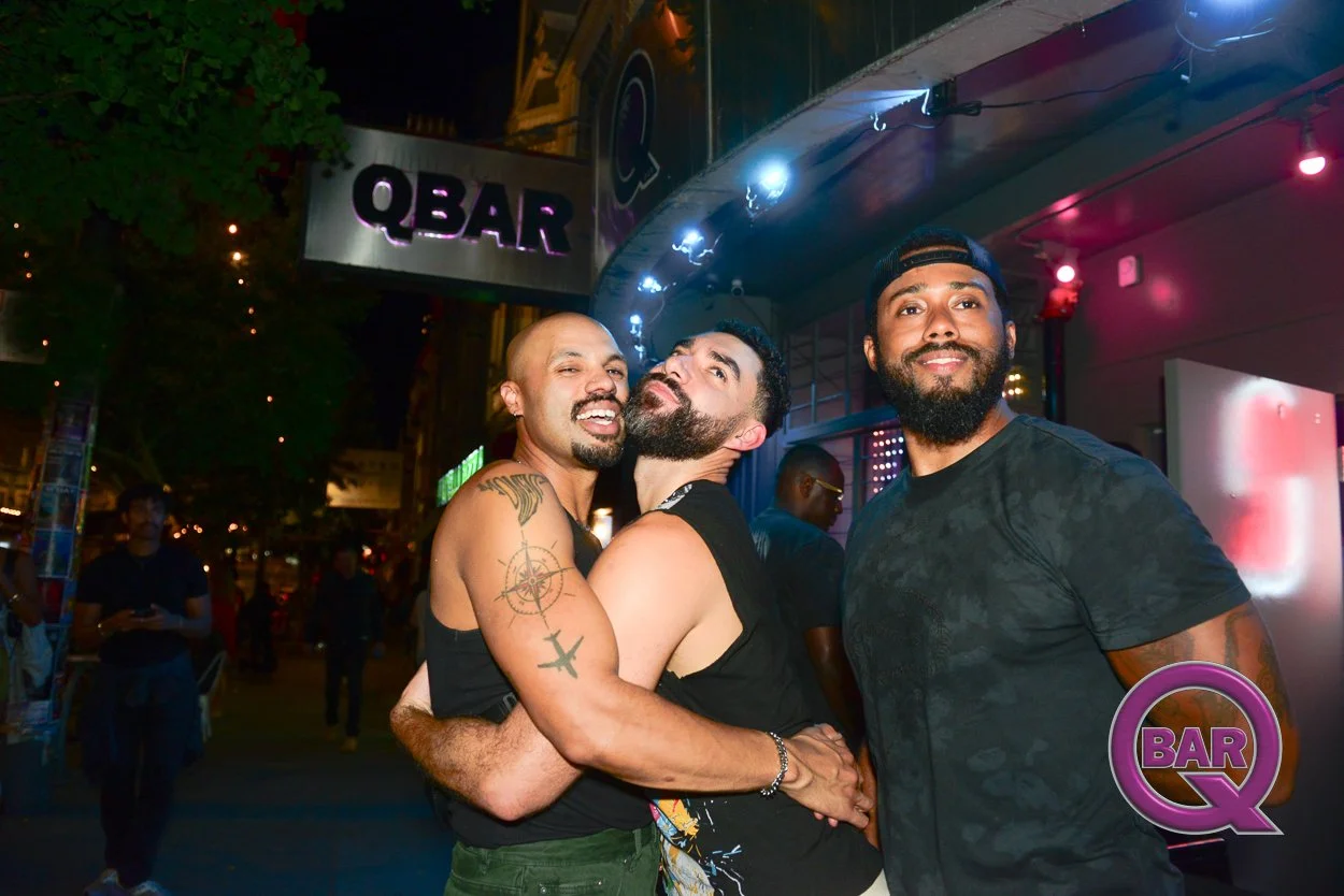 Three men smiling and hugging outside Q Bar at night, with colorful neon lights and a group of people in the background.