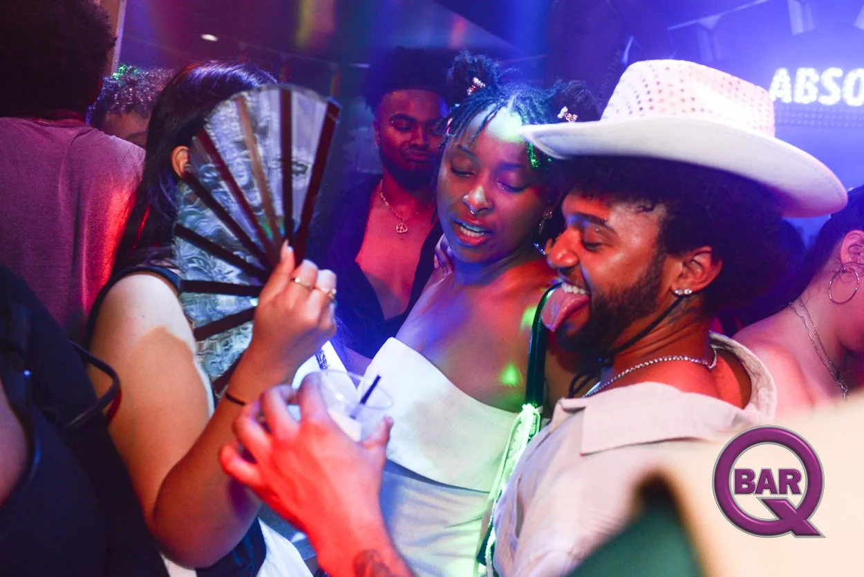 People dancing and having fun at a lively nightclub or bar, with colorful lights and a neon sign in the background, including a woman holding a fan and drinking from a glass with a straw, and a man wearing a white hat sticking out his tongue.