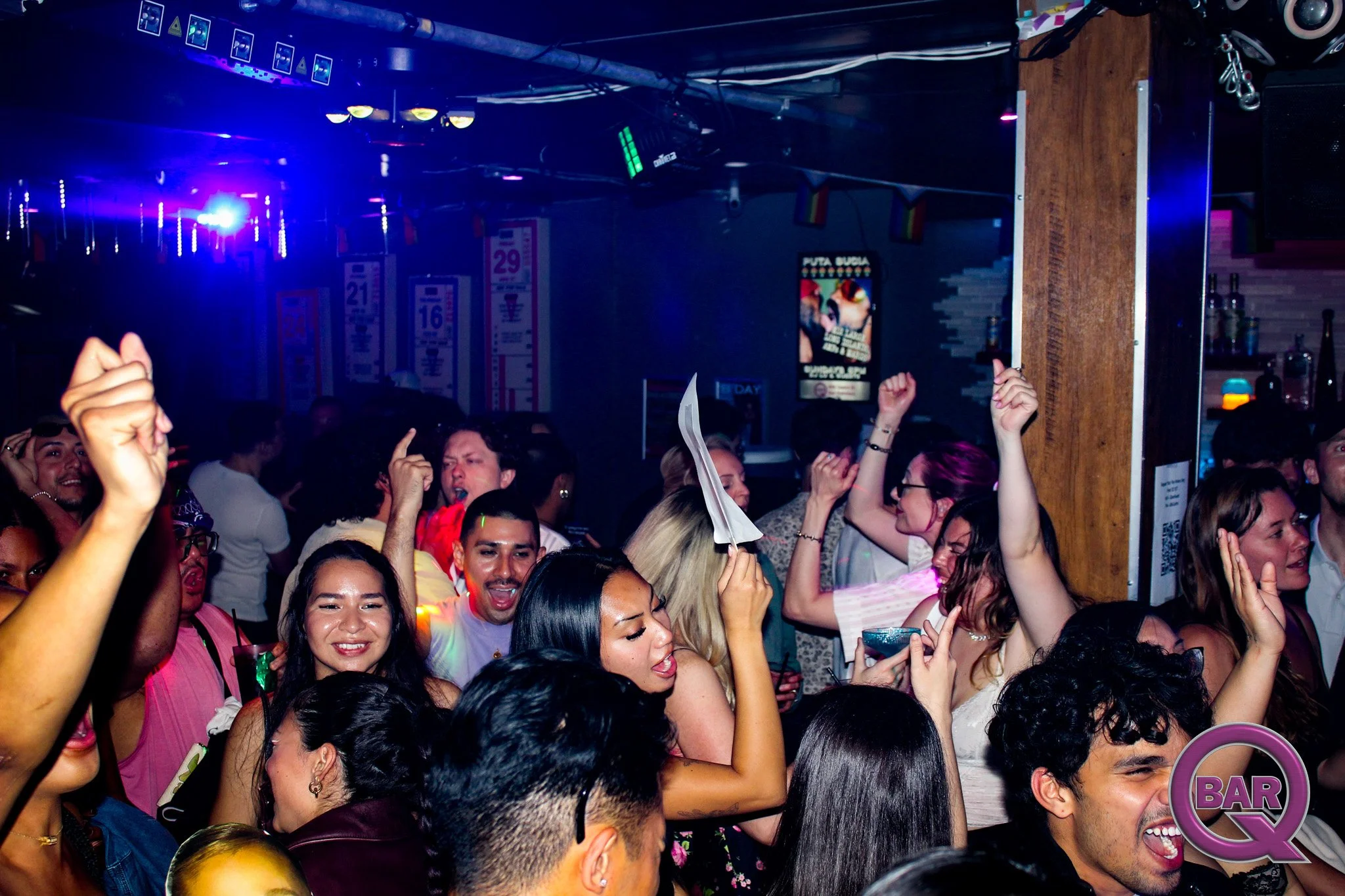 Crowd of people dancing and celebrating at a nightclub with colorful lighting and a DJ booth in the background.