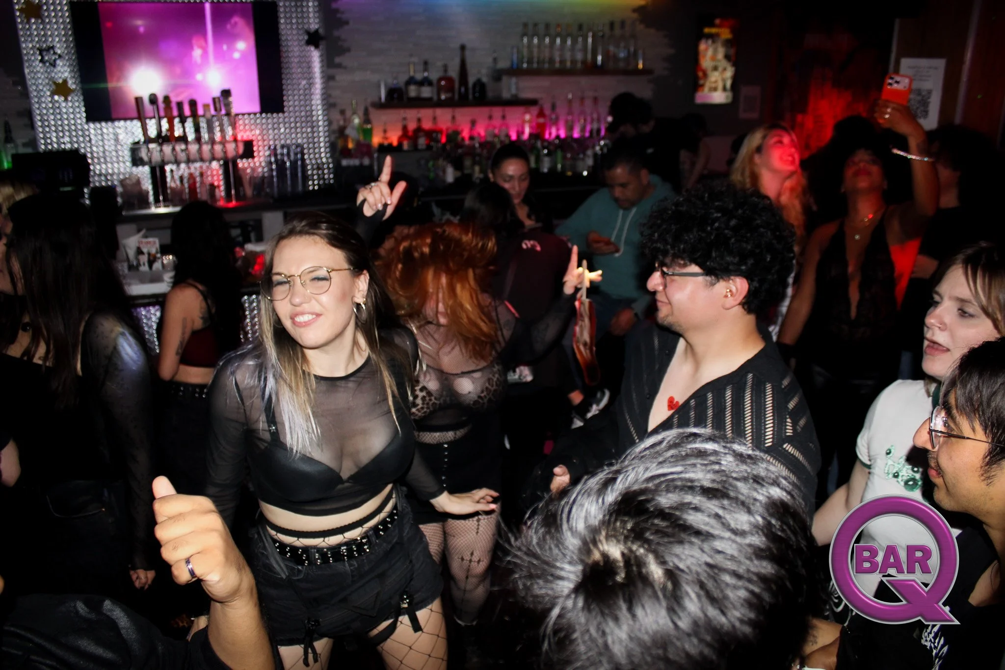 People dancing and socializing in a dimly lit nightclub with a pink-lit bar in the background and a screen displaying visuals.