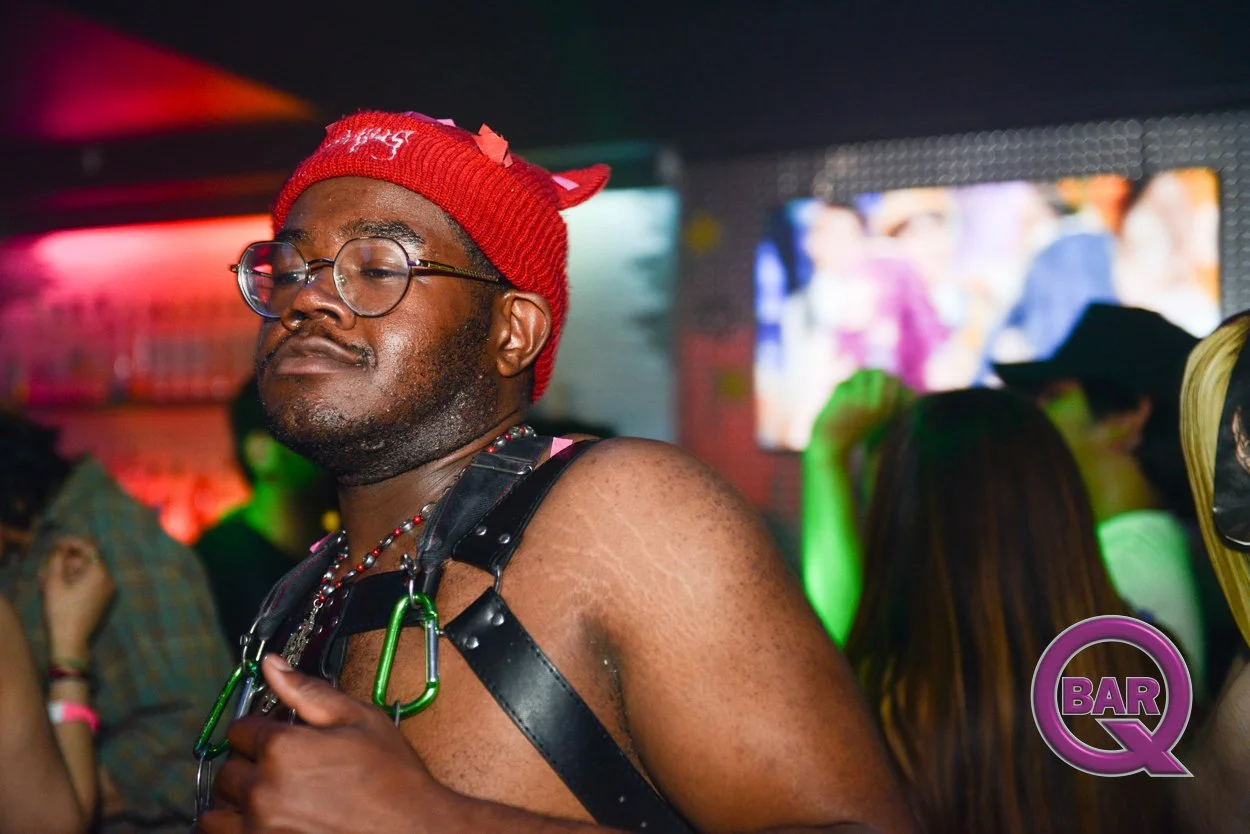 A man wearing glasses, a red knit cap, and a black leather harness with green clips, standing in a dimly lit environment with colorful lights, possibly a nightclub or bar. In the background, other people are dancing or socializing, and a TV screen is