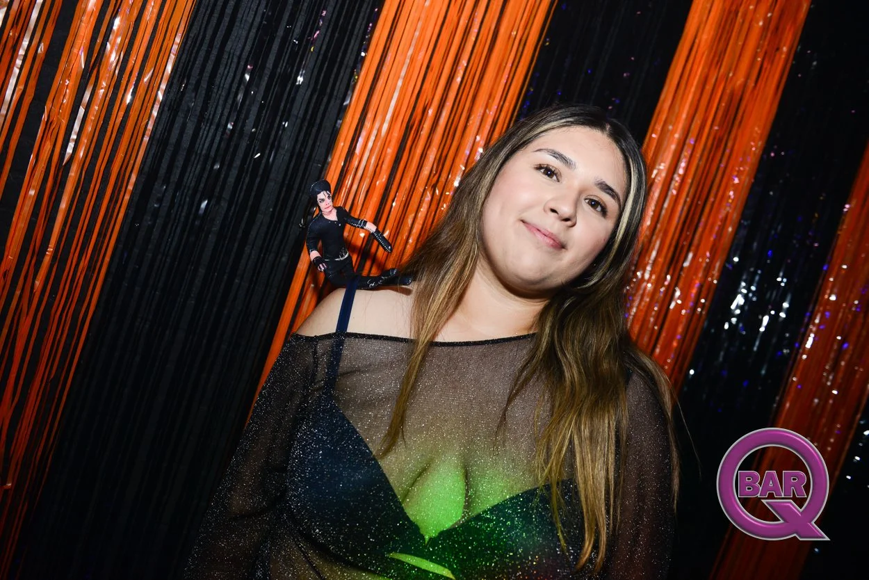A woman with long brown hair smiling in front of black and orange streamers at a party or bar, with a Barbie doll on her shoulder. There is a logo in the lower right corner that says 'Bar Q'.