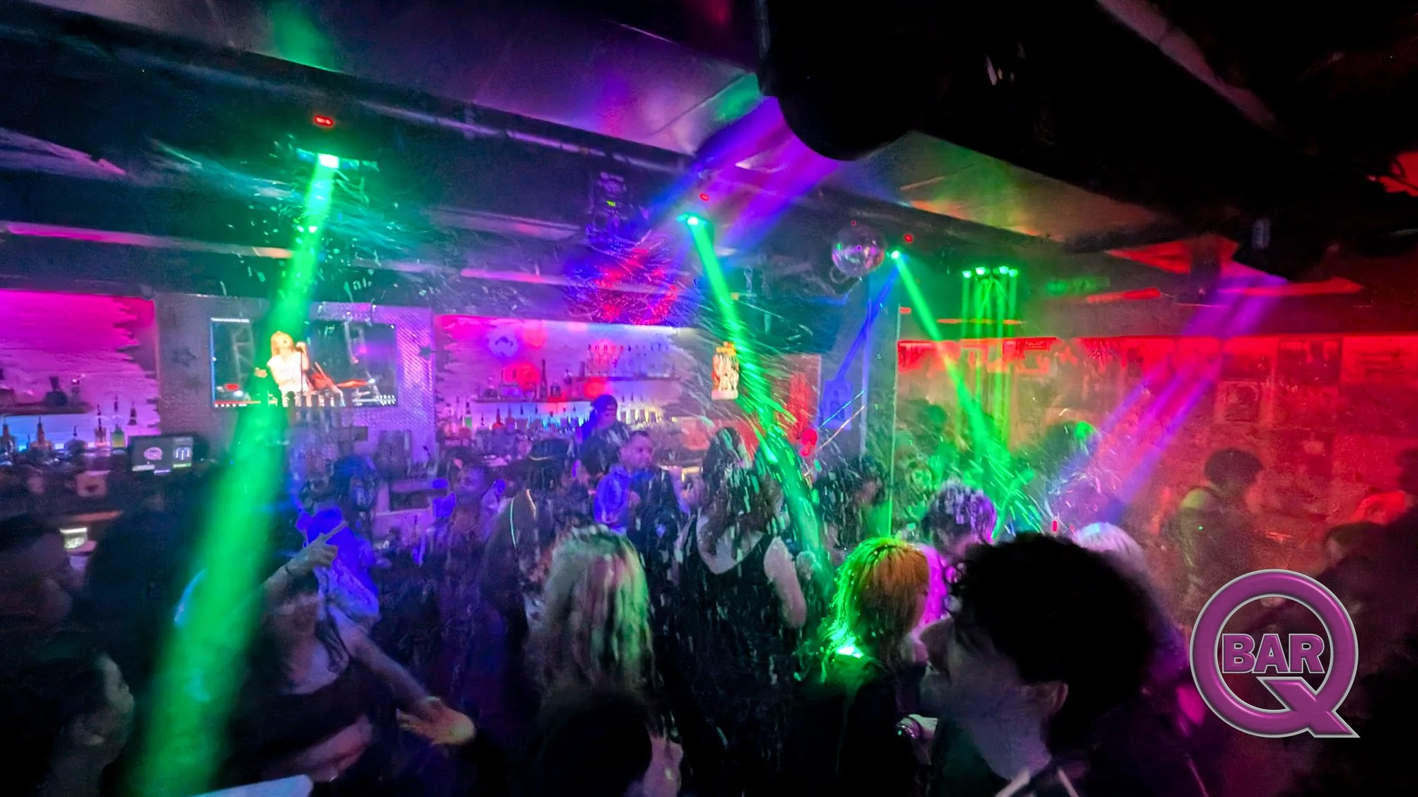 A crowded nightclub with colorful laser lights and a bar area, people dancing and socializing under vibrant lighting.