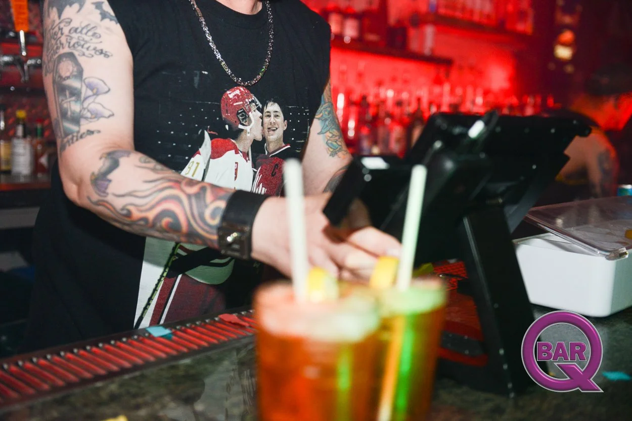 A bartender with tattoos on his arms working behind a bar counter with colorful drinks, a cash register, and various bottles in the background. The bartender is wearing a black t-shirt with sports team graphics and a chain necklace, with a prominent 