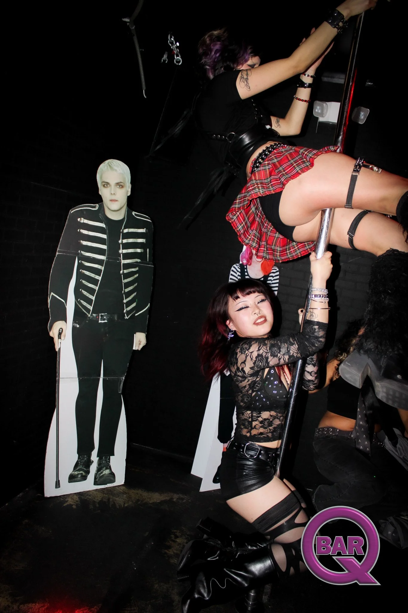 People holding a cutout of a person with platinum blonde hair in a military-style jacket, and a woman in black lingerie, a red plaid skirt, and thigh-high stockings dancing with a pole at a nightclub.