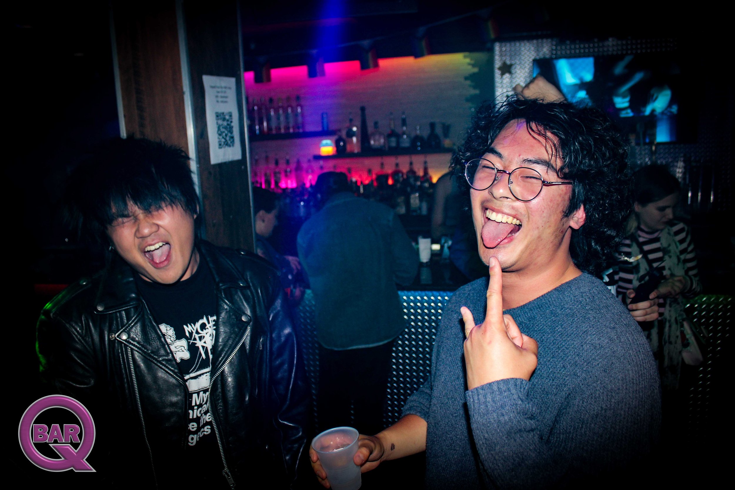 Two young men are smiling and joking at a bar or nightclub with colorful lighting in the background. One man has glasses and curly hair, sticking out his tongue and holding a finger up, while the other man has dark hair and is wearing a black leather
