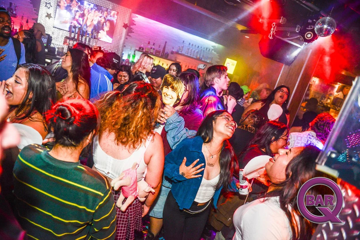 Crowd of people dancing and enjoying themselves at a lively bar or nightclub with colorful lights, a large screen in the background, and a bar area with bottles and glasses.