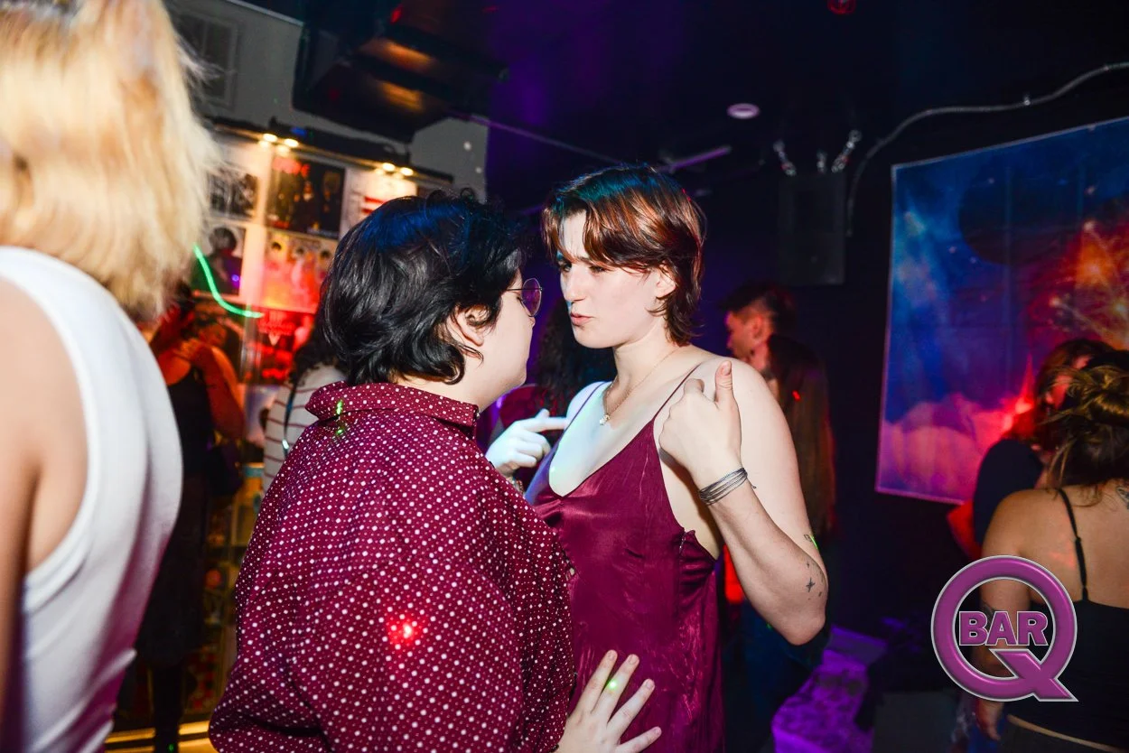 Two women dancing and talking in a nightclub with colorful lights and other people in the background.