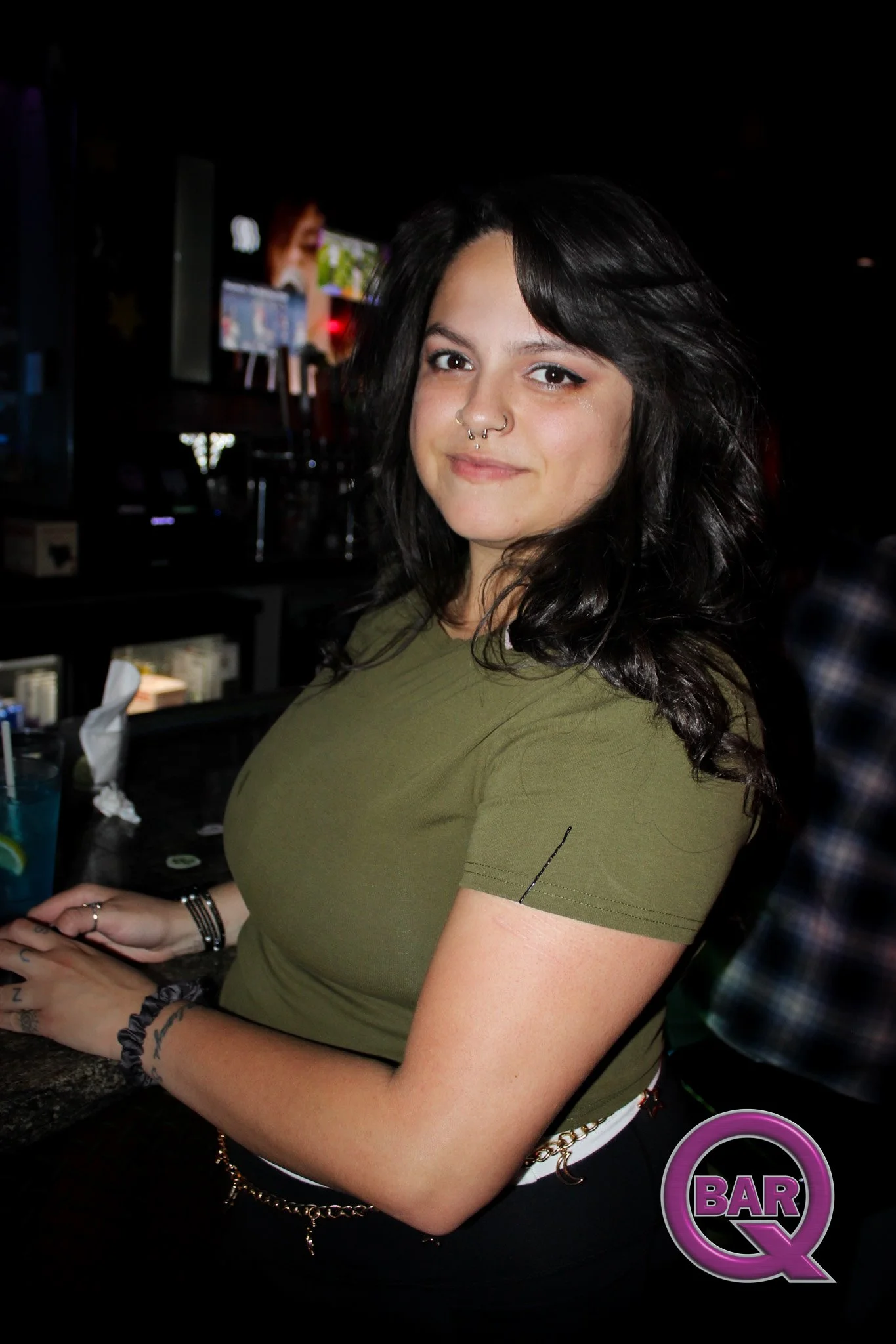 A young woman with dark hair, pierced nose, and tattoos on her arm, standing at a bar with a purple neon sign that says 'BAR Q', in a dimly lit setting.