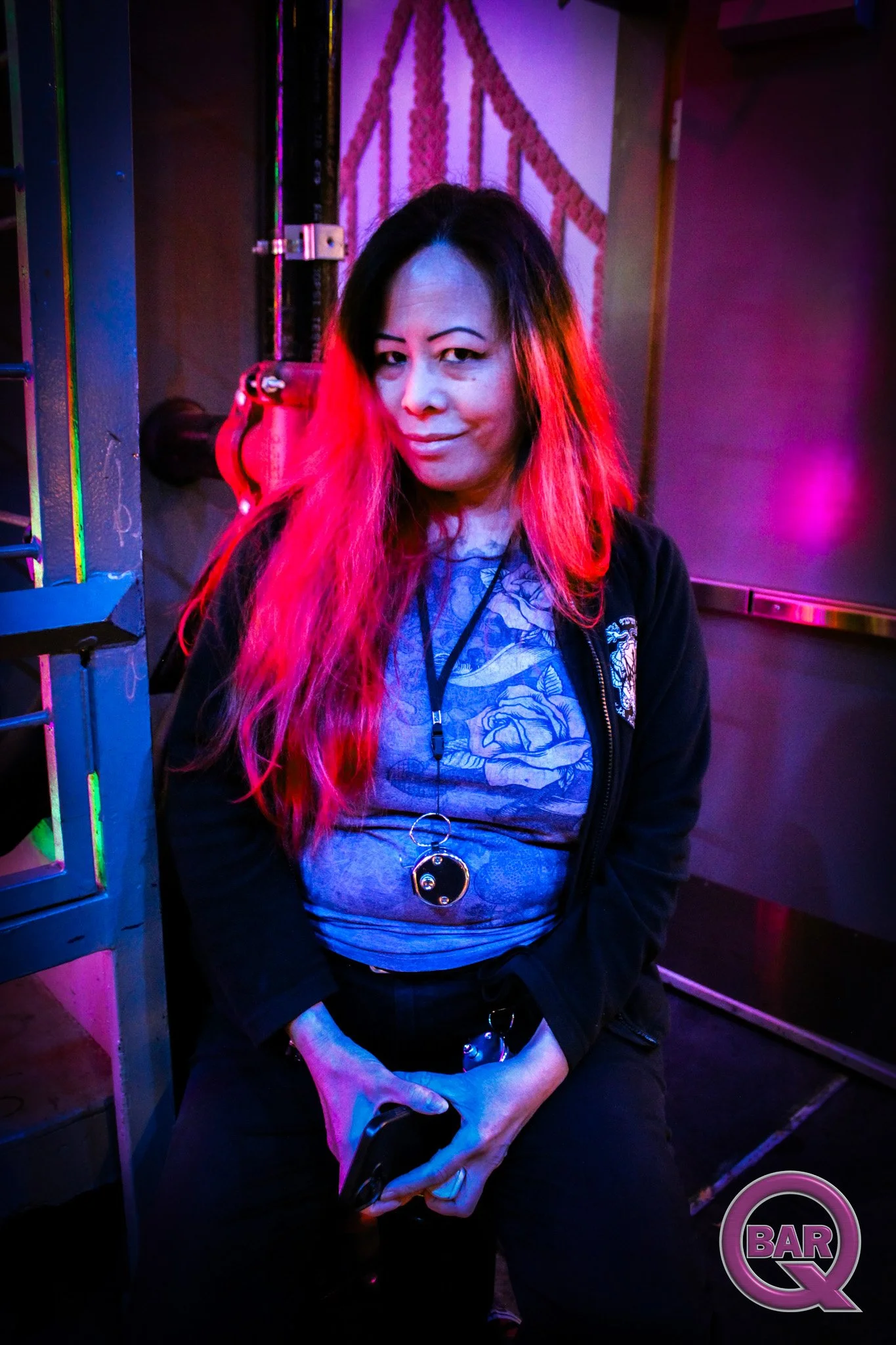 A woman with long red hair, wearing a black jacket and a blue shirt with a floral design, holding a black object in her lap, sitting in a dimly lit bar with colorful neon lights and a bar sign in the background.